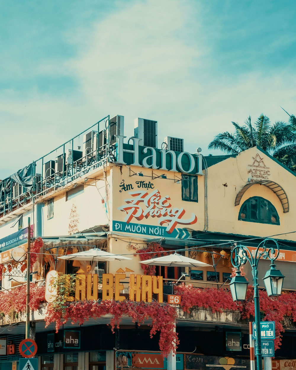 Two-story building corner with a yellow facade covered in red vines and umbrellas over outdoor seating, featuring large signs with Vietnamese text and 'Hanoi' on top, under a partly cloudy blue sky with palm trees on the side.