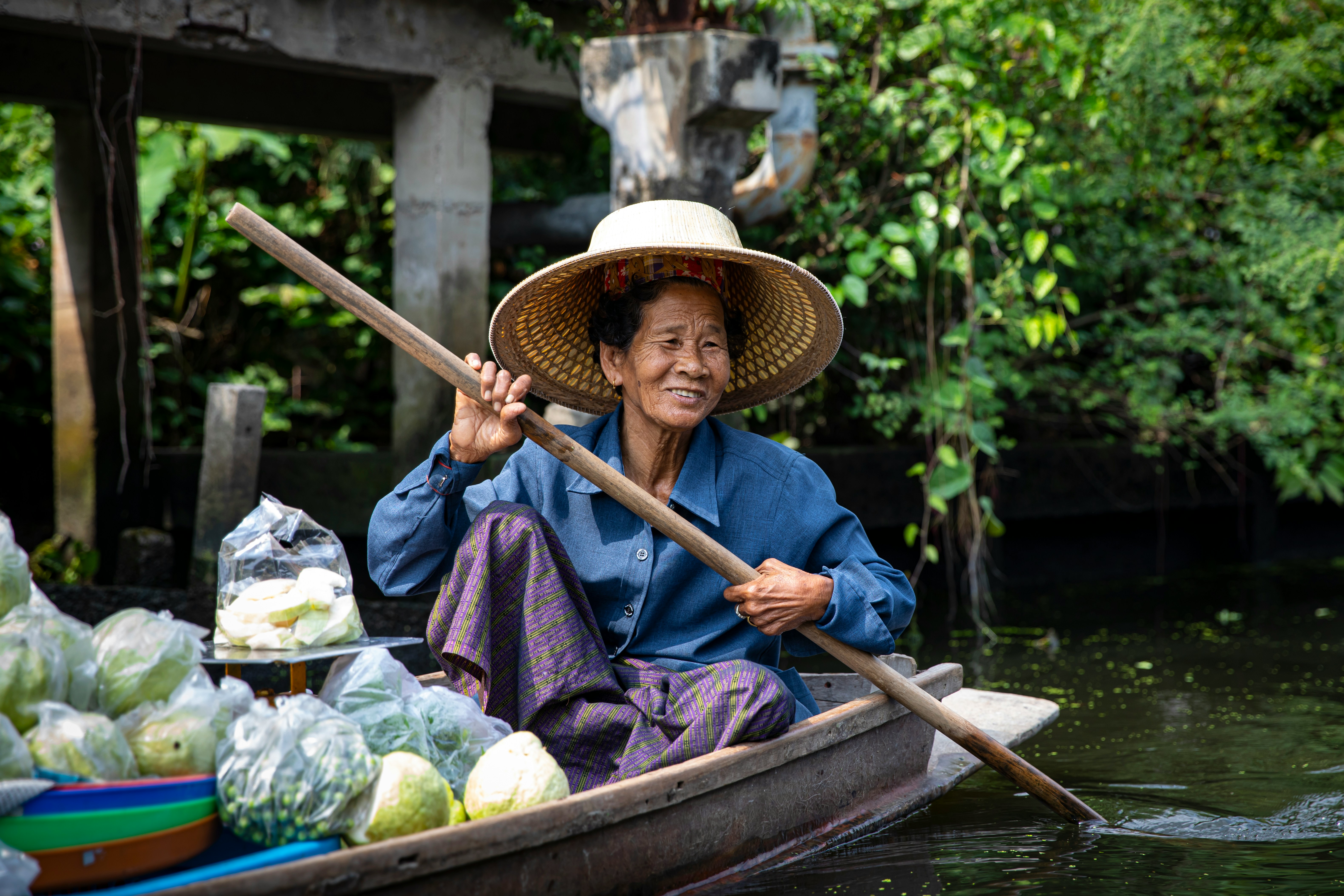 Smiling elderly woman wearing a wide-brimmed straw hat and blue shirt paddling a wooden boat filled with plastic-wrapped vegetables on calm water, with green foliage in the background.