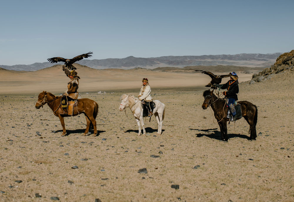 Three riders on horseback in a rocky, dry plain with mountainous background; two riders hold large eagles perched on their gloved arms, and the middle rider sits on a white horse without an eagle.