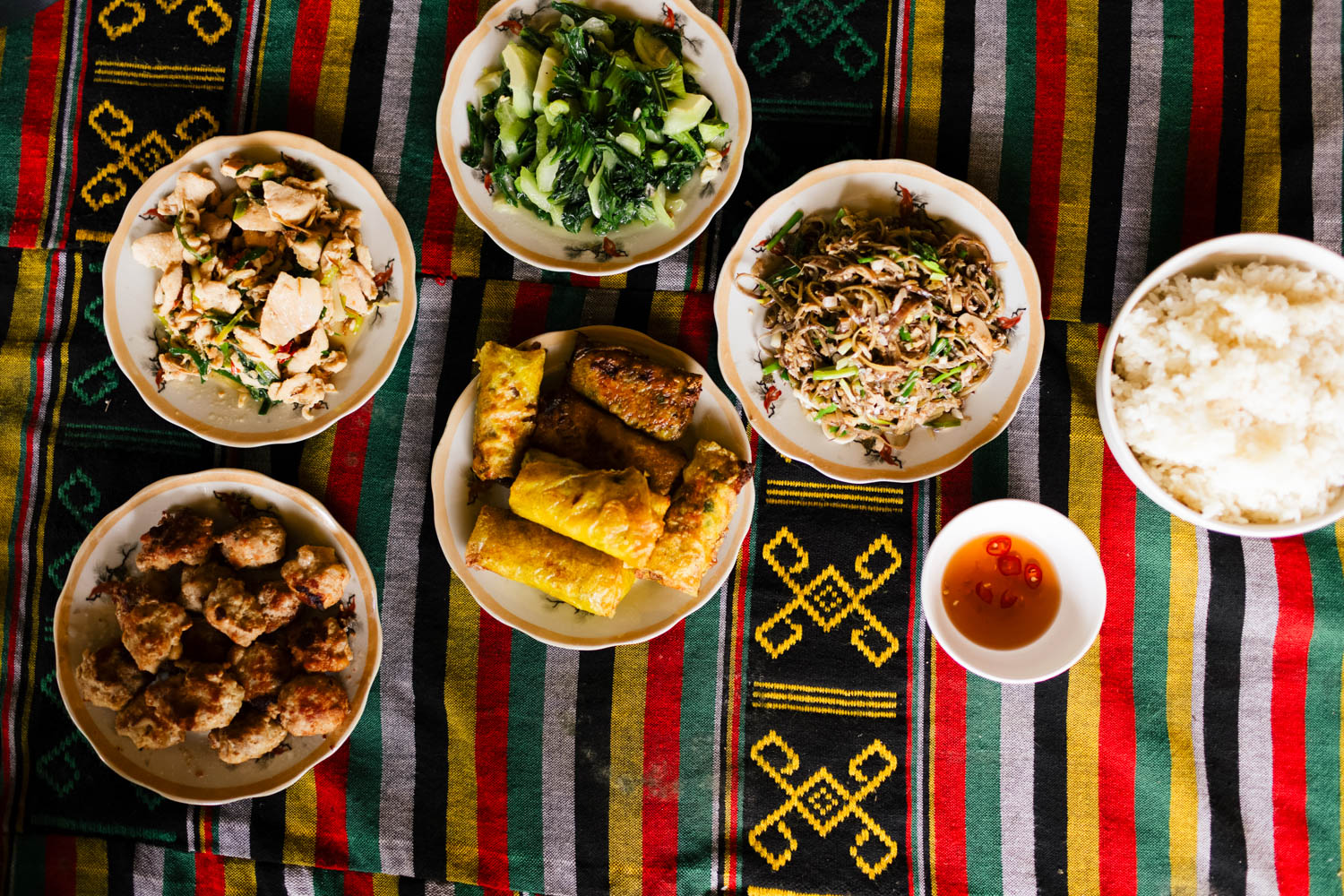 Top-down view of six plates with various Asian dishes on a colorful striped and patterned cloth: stir-fried chicken with vegetables, cooked leafy greens, stir-fried noodles with shredded meat, fried spring rolls, fried meatballs, a bowl of white rice, and a small bowl of chili sauce.