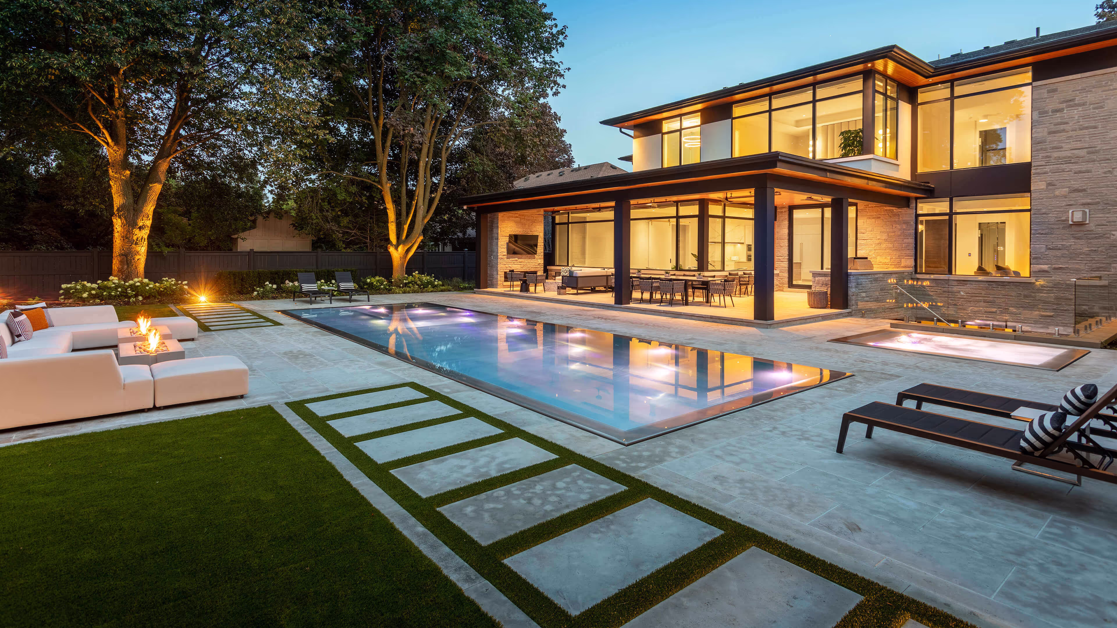 Modern two-story house with large windows, illuminated outdoor patio, swimming pool, hot tub, and seating area with fire pit at dusk.