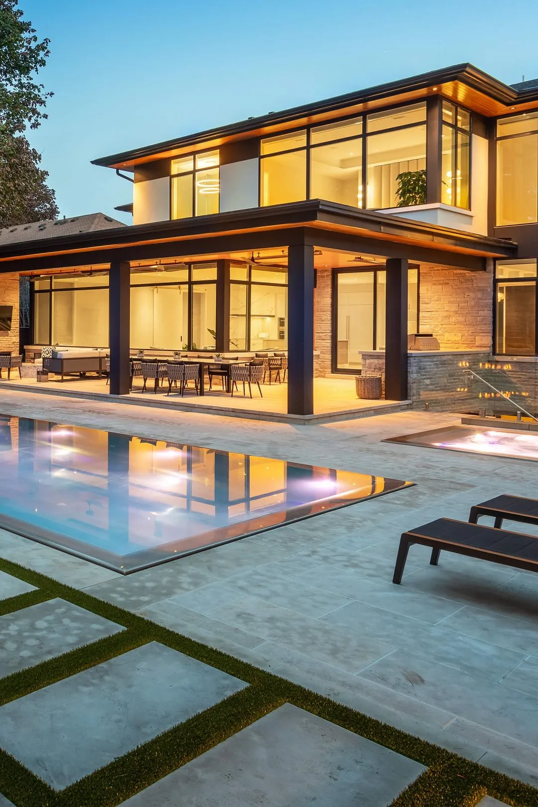 Modern two-story house with large glass windows, outdoor dining area, swimming pool, and patio lounge chairs at dusk.