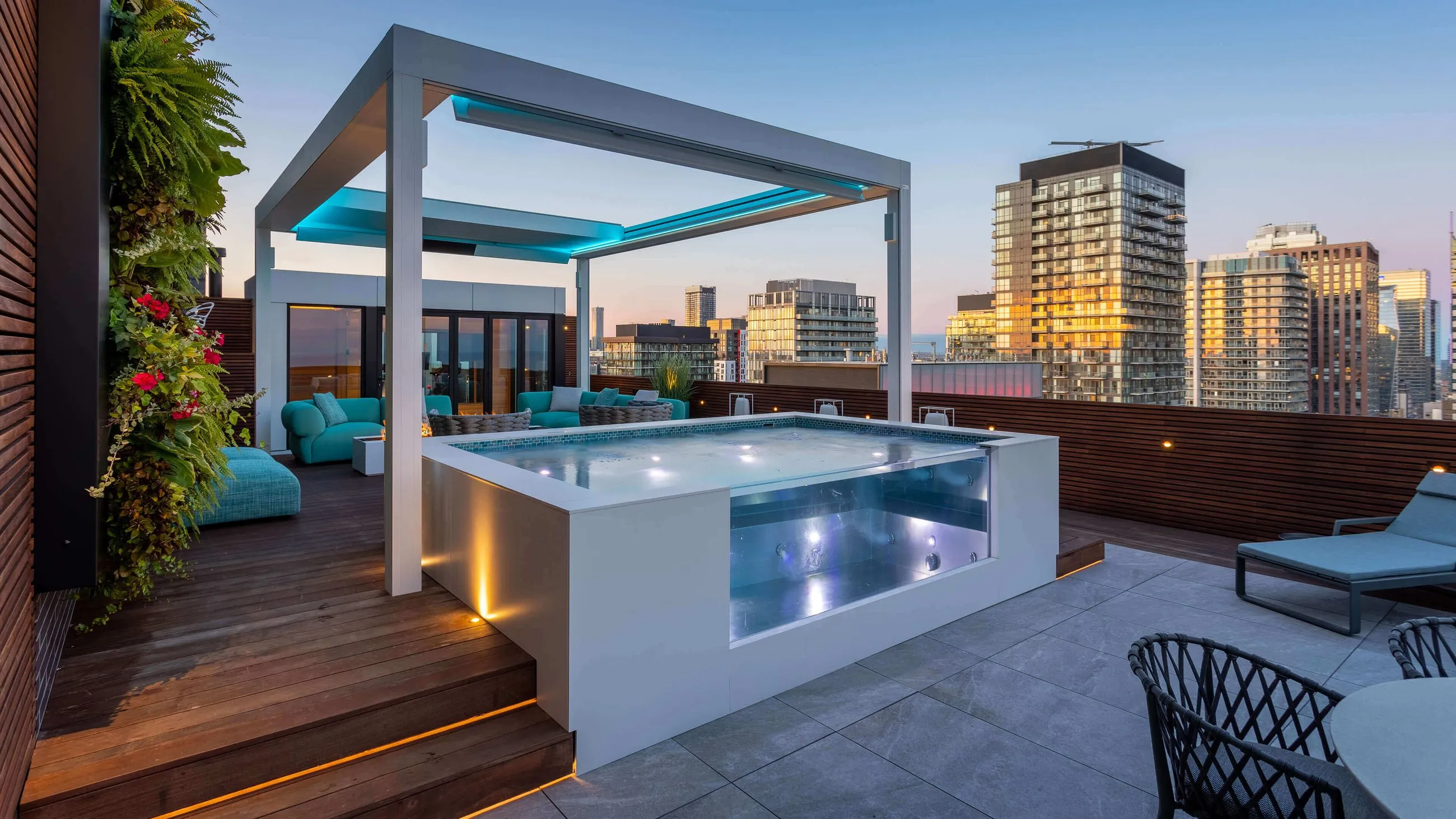 Modern rooftop terrace with a lit white jacuzzi, teal lounge furniture, vertical garden, and city skyline at sunset.
