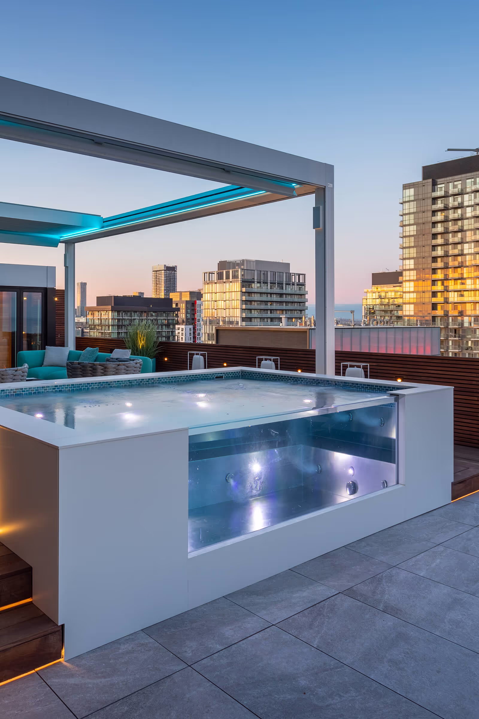 Modern rooftop patio with a glass-fronted hot tub, seating area, and city buildings at sunset.
