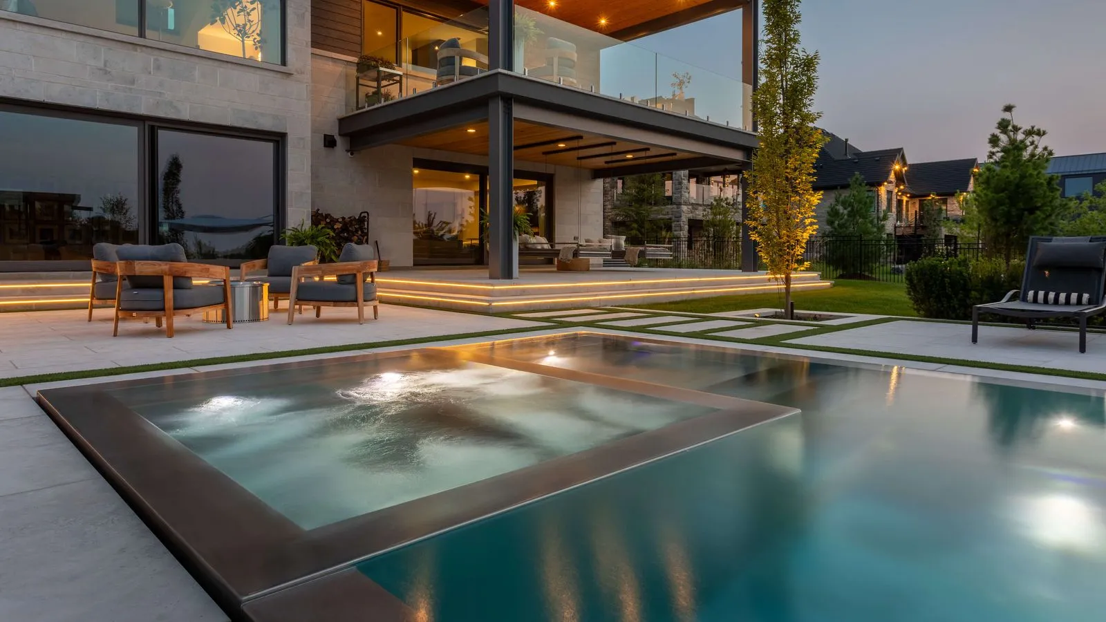 Modern home with a lit patio, wooden chairs, and a spa pool adjacent to a swimming pool at dusk.