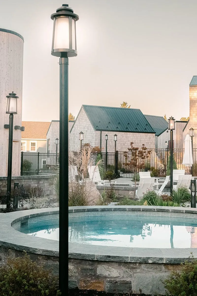 Outdoor stone hot tub surrounded by lampposts and white chairs with buildings in the background at dusk.