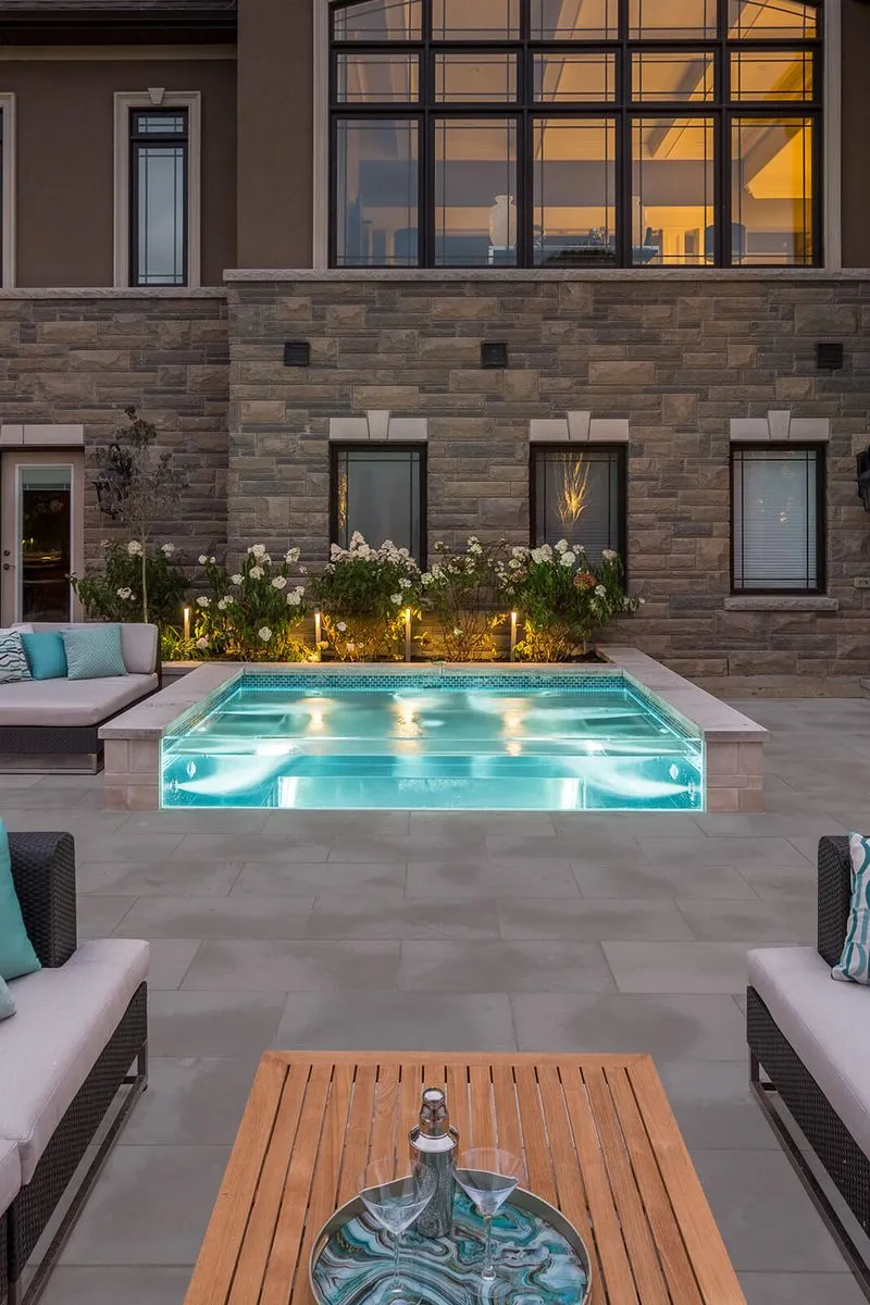 Transparent outdoor pool with illuminated water surrounded by patio seating and stone building.