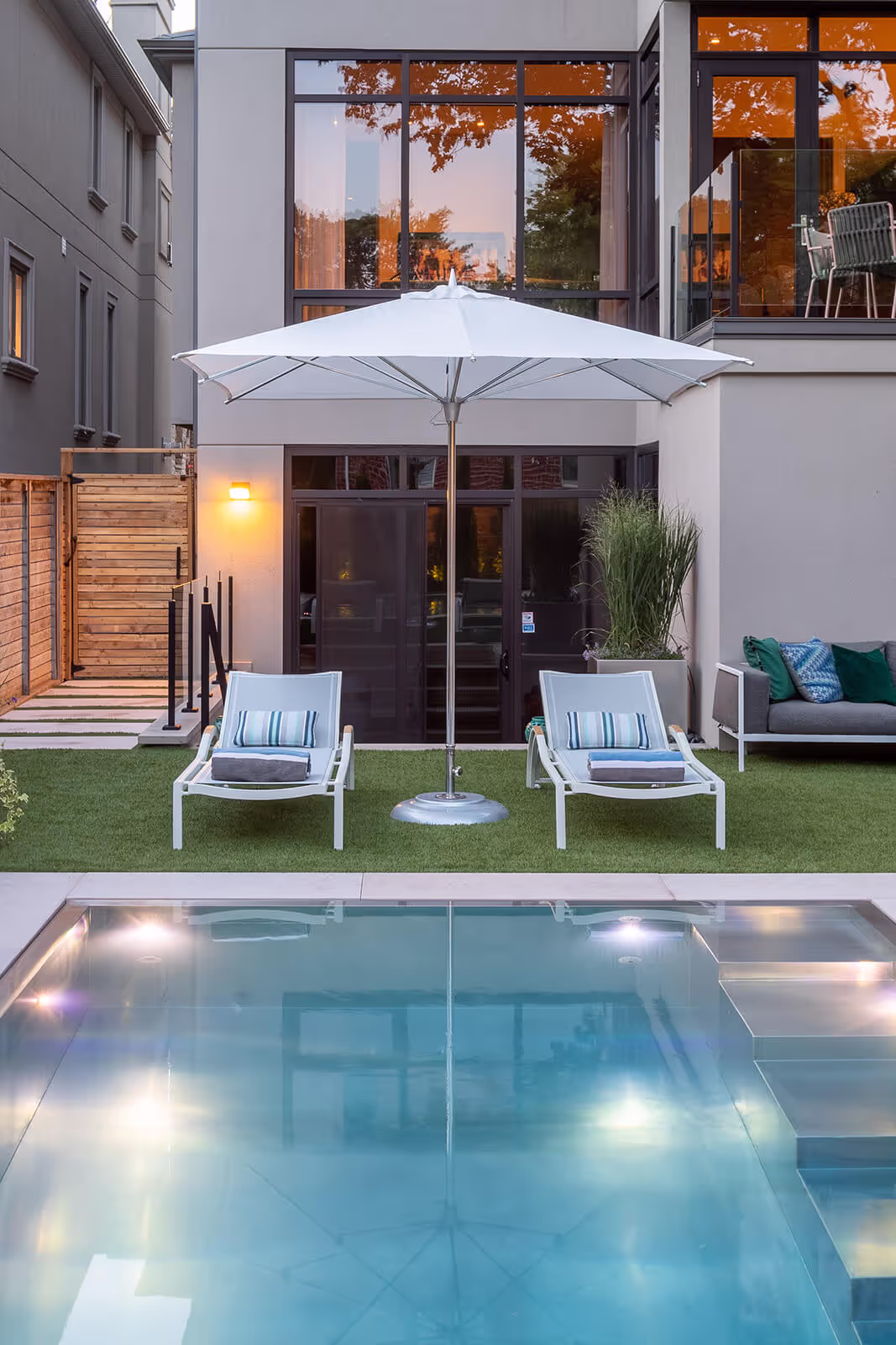 Modern backyard with swimming pool, two lounge chairs under a large white umbrella, and a gray sofa with cushions.