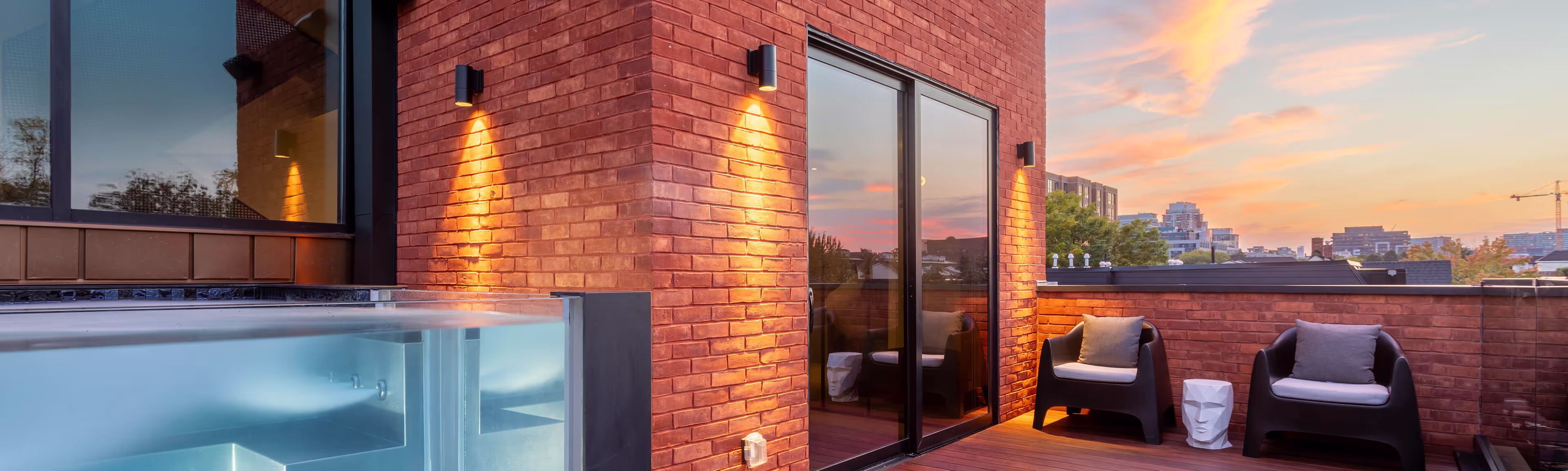 Modern rooftop terrace with glass-walled hot tub, two black chairs with cushions, white sculptural side table, and city skyline at sunset.