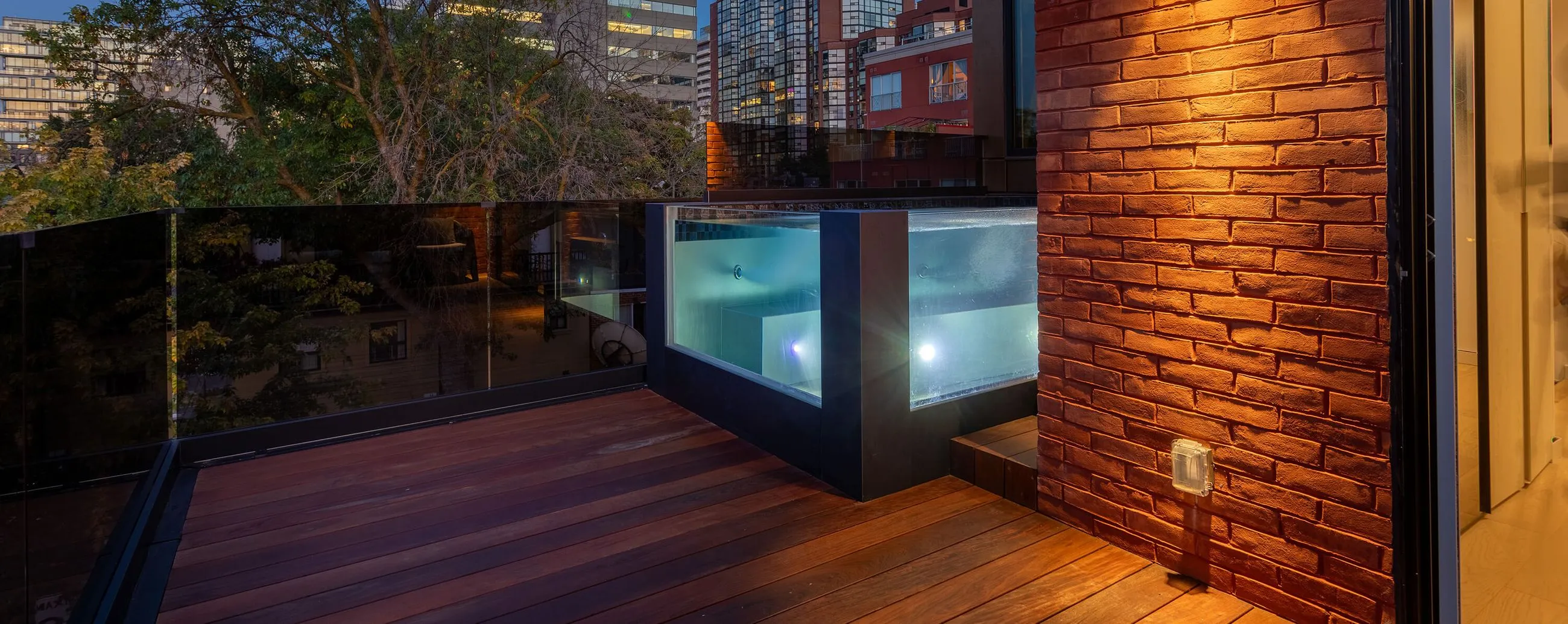 Modern rooftop deck with wooden flooring, glass railing, and illuminated built-in hot tub next to brick wall at dusk.