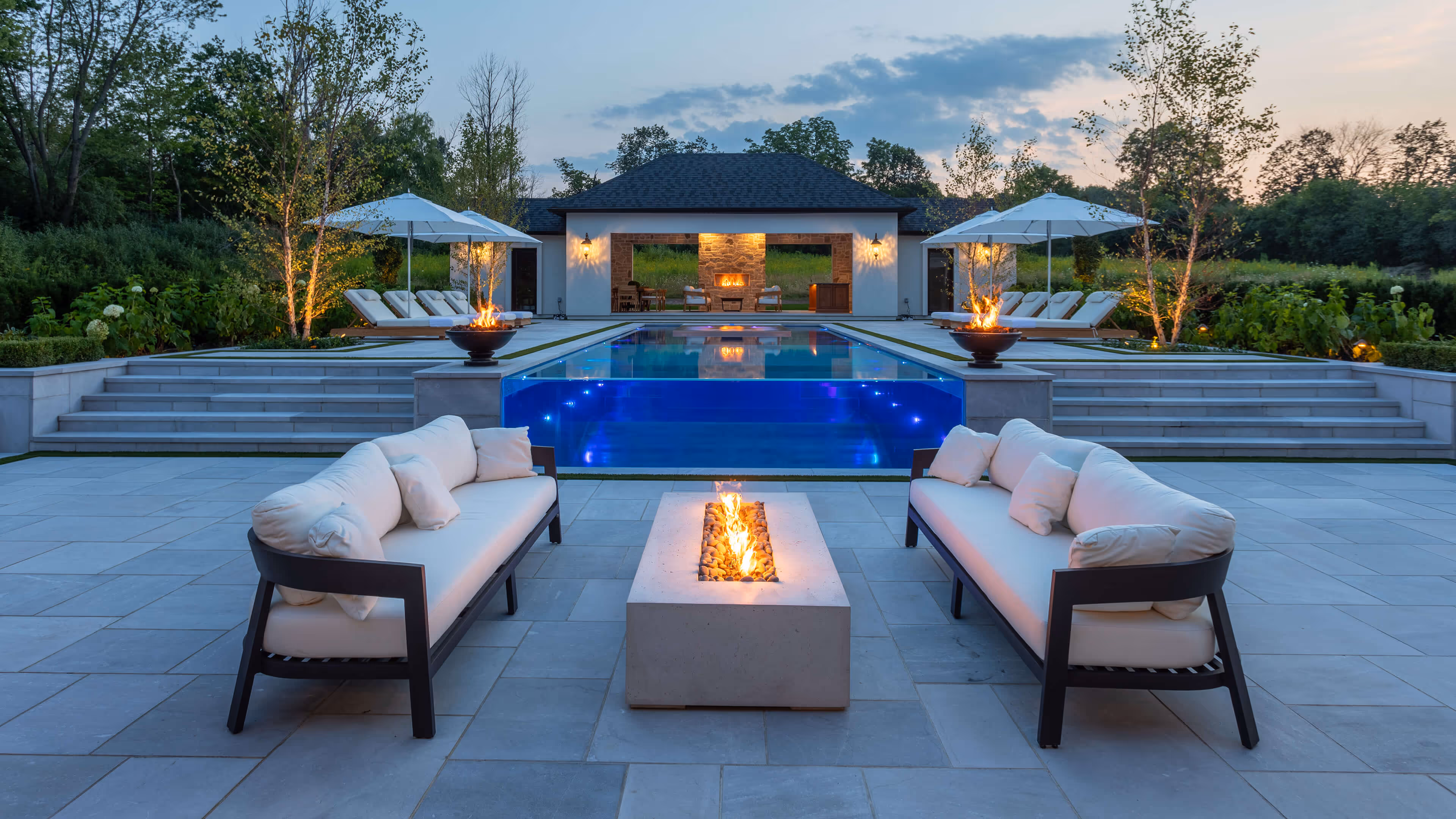 Modern outdoor patio with two white cushioned sofas facing a rectangular fire pit, overlooking an illuminated infinity pool and a pool house with a fireplace.