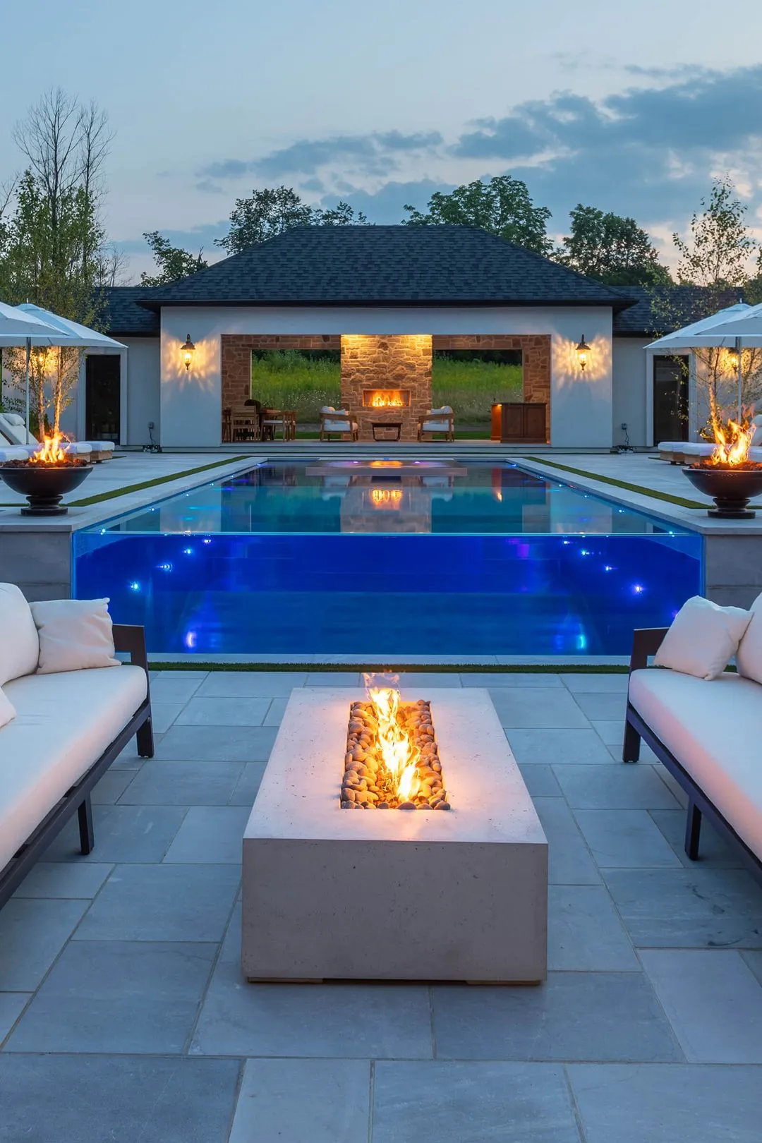 Modern backyard with a rectangular glass-edged pool, fire pit between two white cushioned sofas, and a lit outdoor fireplace in the pavilion at dusk.