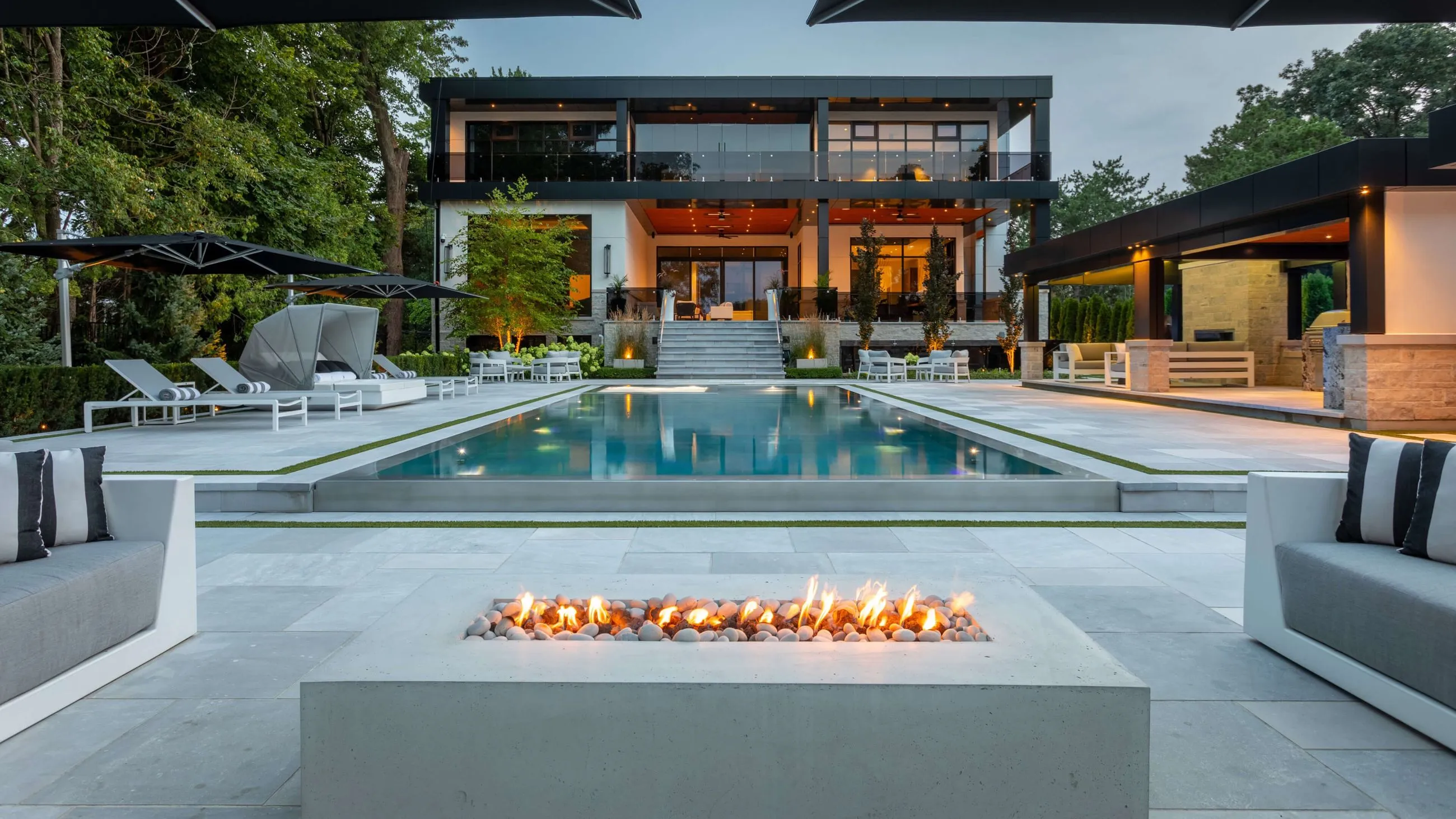 Modern two-story house with a large pool, outdoor fire pit, and patio lounge area surrounded by trees.