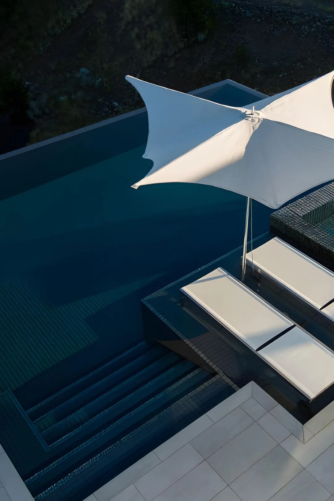 A modern poolside area with two white lounge chairs under a large white umbrella beside clear blue water and steps leading into the pool.