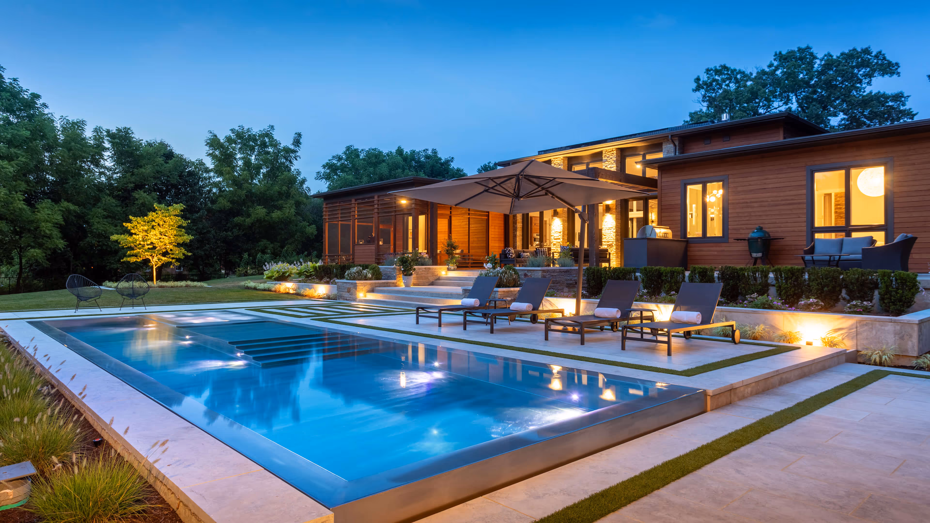 Modern backyard with illuminated swimming pool, loungers under umbrella, and a lit house at dusk.
