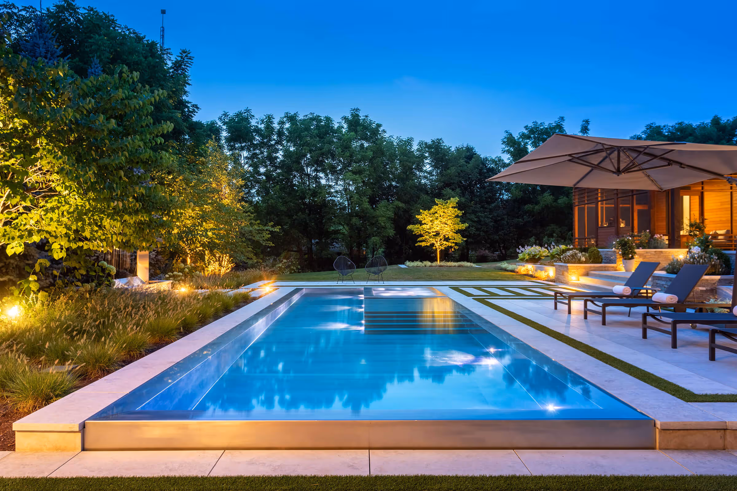 Modern backyard pool at dusk with lounge chairs, umbrella, and garden lighting around green trees.