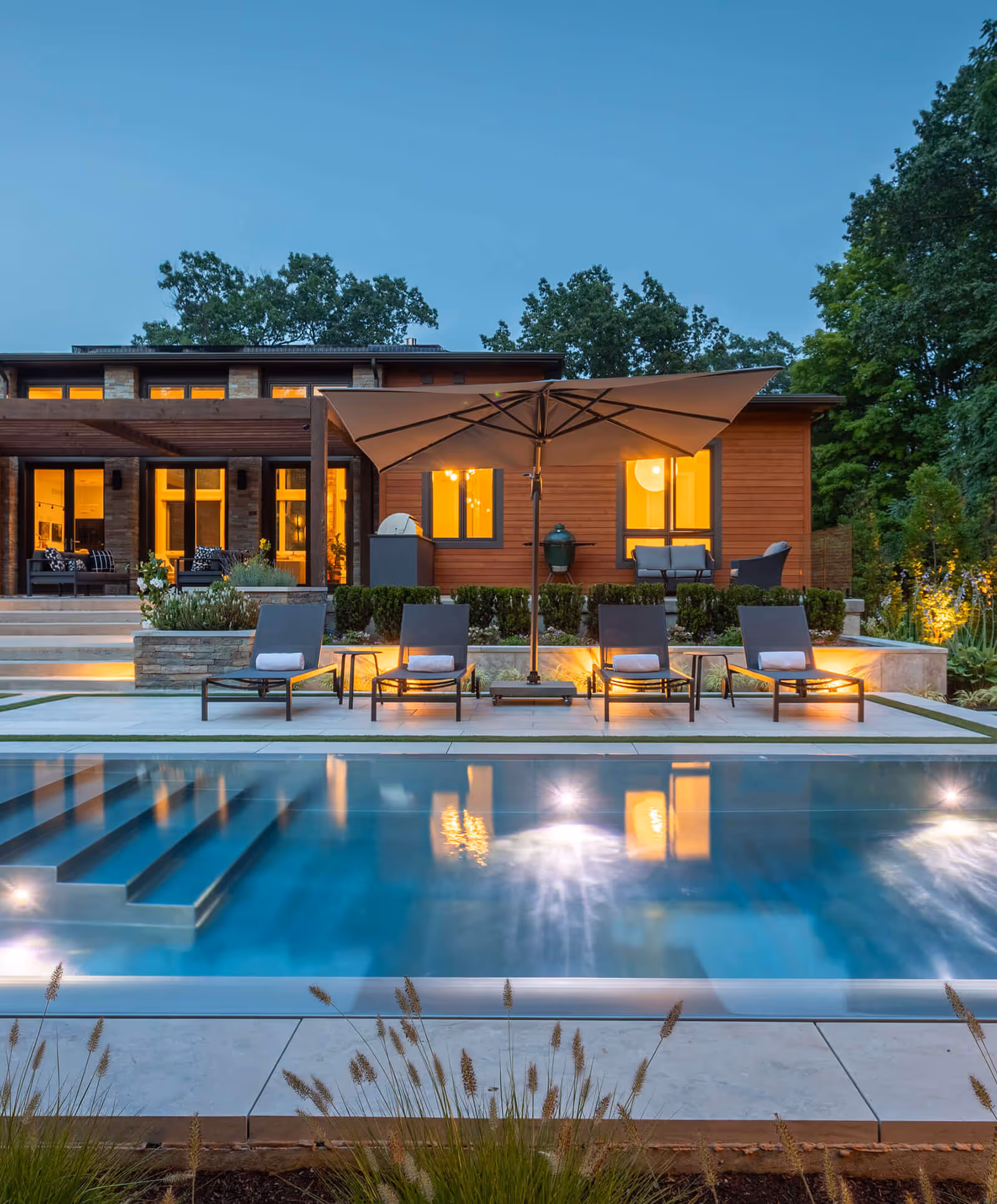 Modern backyard with lit pool, four lounge chairs under large umbrella, and house with glowing windows.