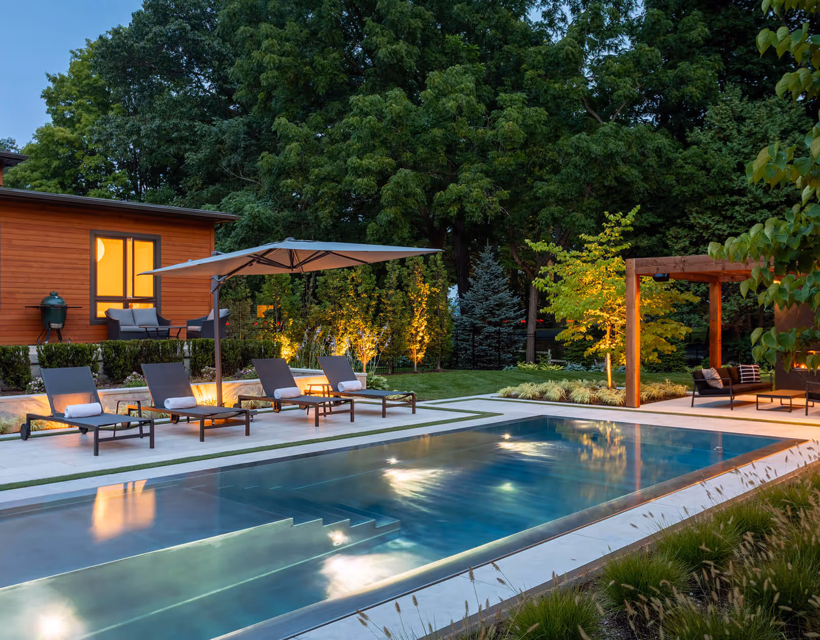 Modern backyard with lit swimming pool, four lounge chairs under umbrella, and a wooden pergola seating area.