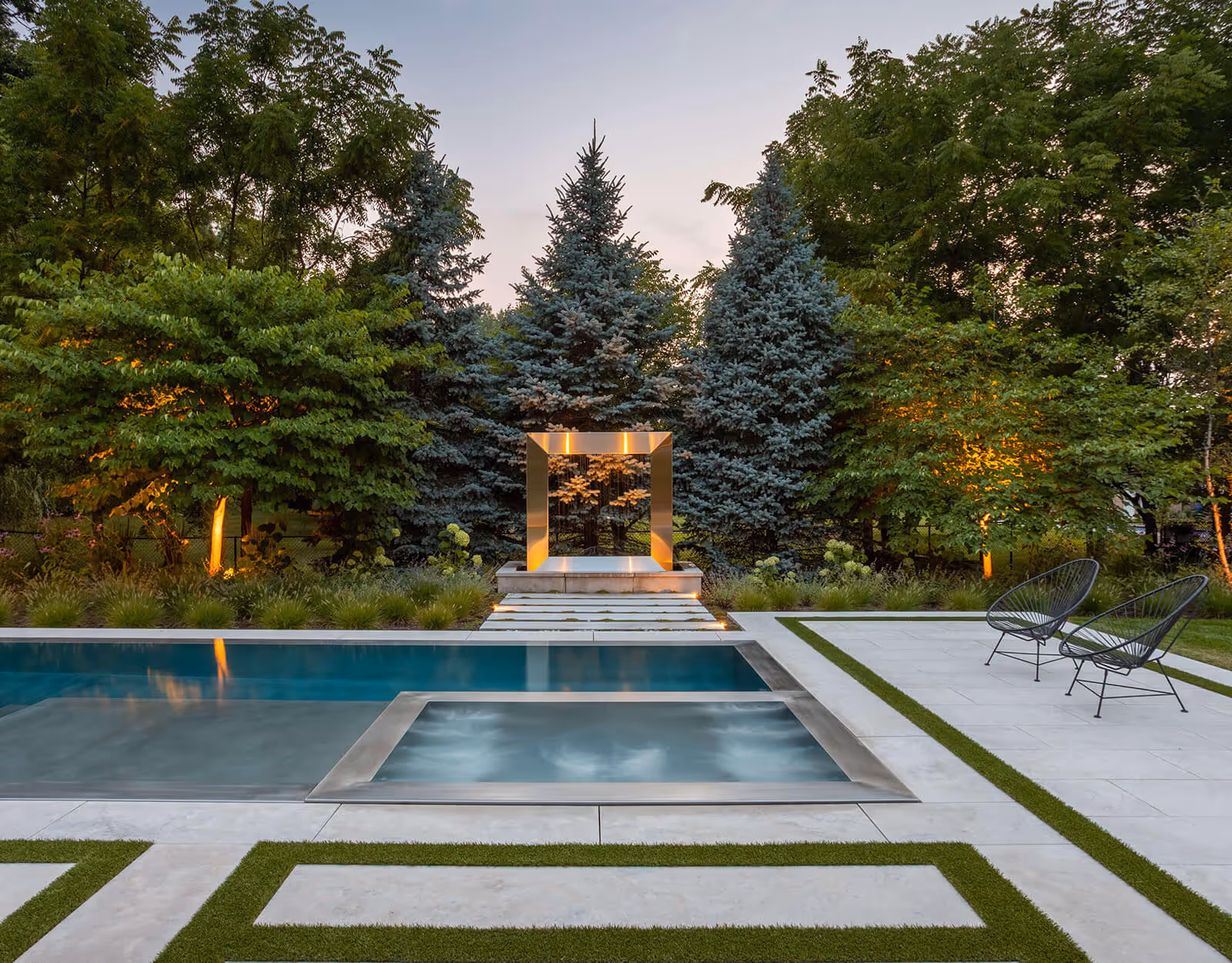 Modern pool and hot tub with geometric tiles, two chairs, and trees lit by outdoor lights at dusk.