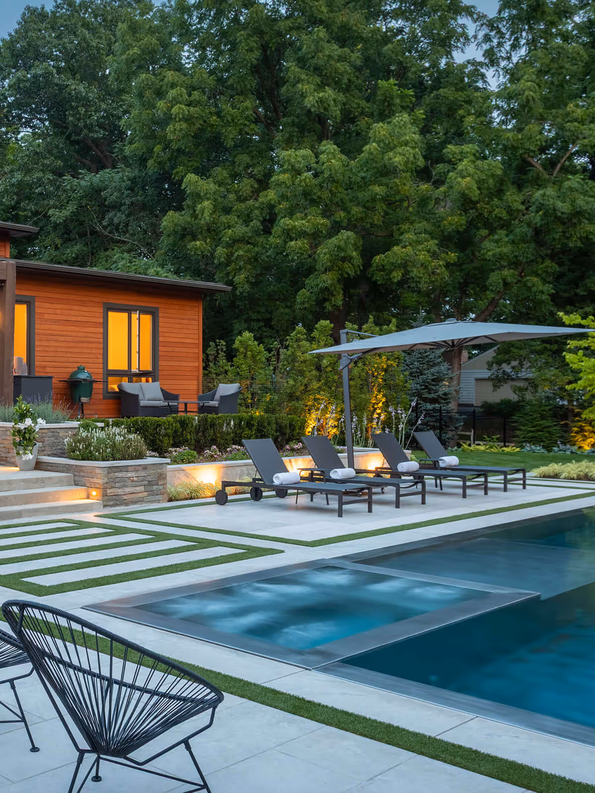 Modern backyard with pool, jacuzzi, four lounge chairs under umbrella, and wooden house with lit windows.