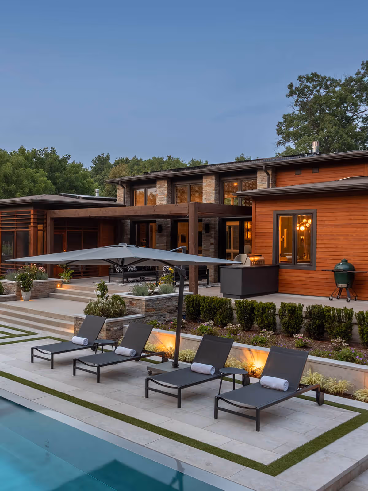 Modern backyard with swimming pool, four lounge chairs under large umbrella, and a wooden house in the back.