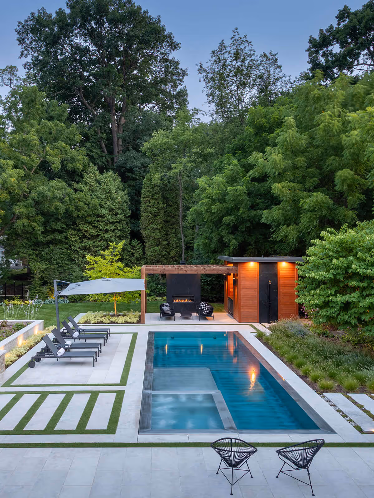 Modern backyard with rectangular pool, lounge chairs, pergola, fireplace, and wood pool house surrounded by trees.