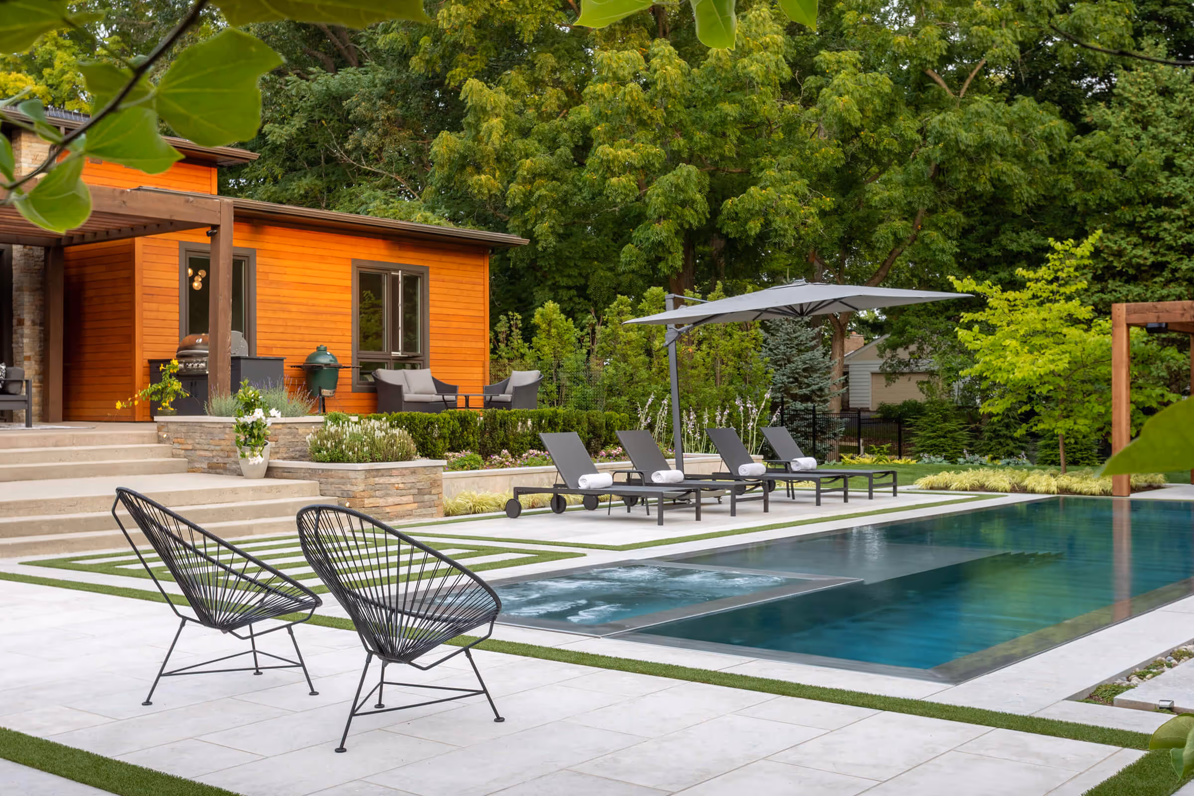 Modern backyard with pool, lounge chairs, umbrella, black chairs, and wood-paneled house surrounded by trees.