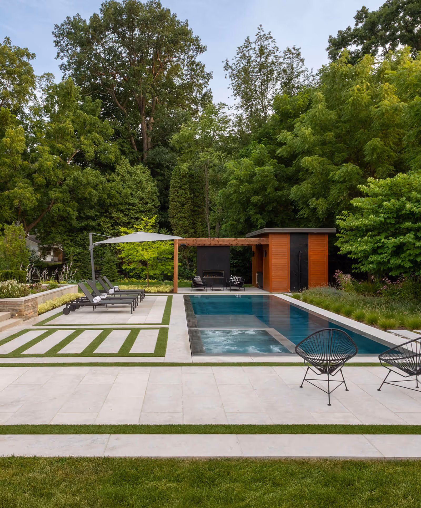 Modern backyard with rectangular pool, lounge chairs, pergola, and lush surrounding trees.