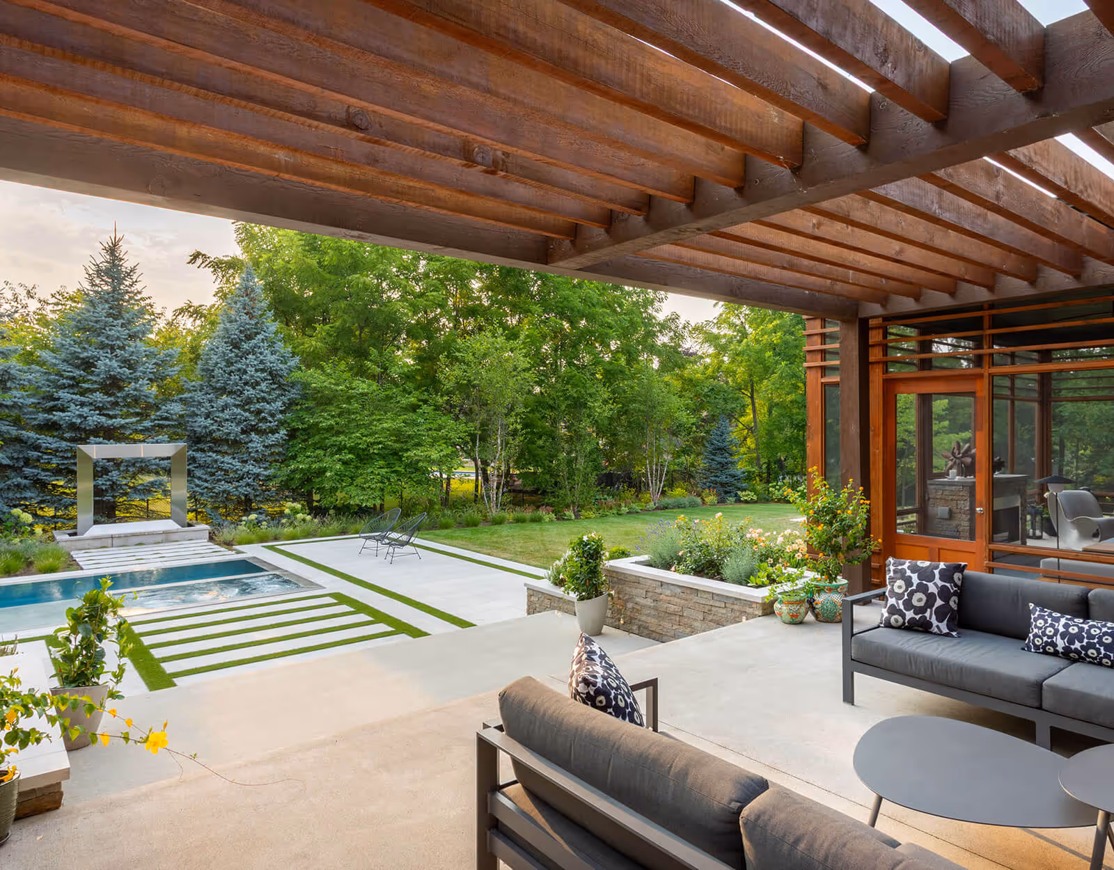 Modern patio with gray sofas under wooden pergola overlooking pool, garden, and green trees.