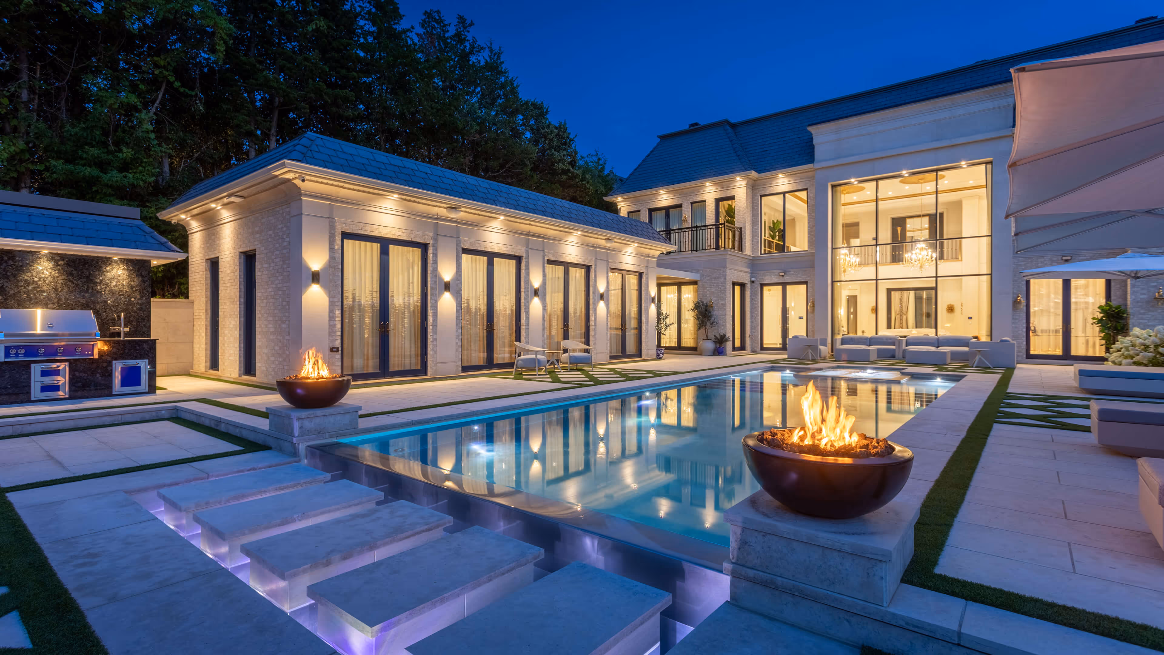Luxurious modern house with illuminated pool, fire bowls, outdoor grill, and patio furniture at dusk.