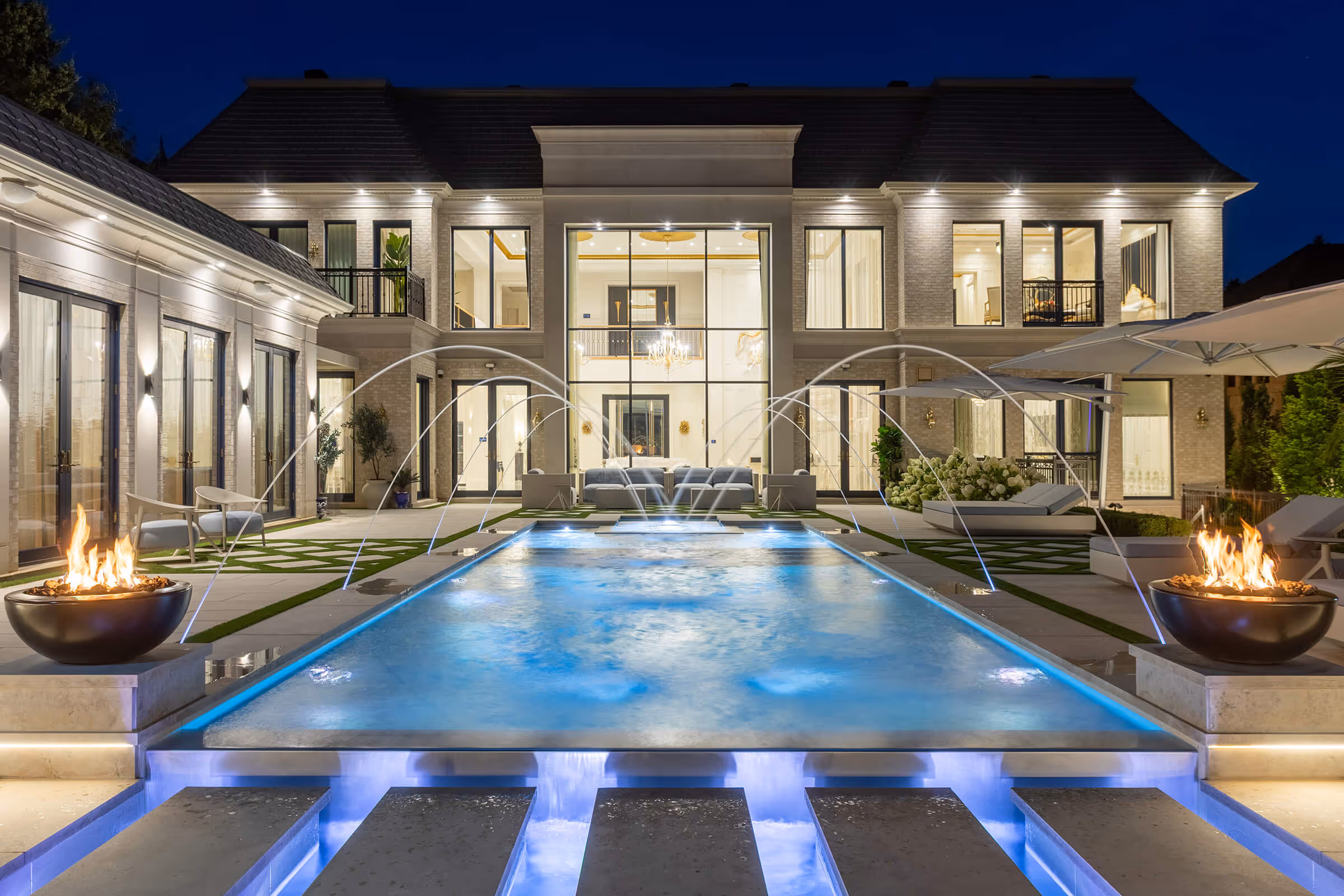 Luxury mansion at night with illuminated pool, water fountains, fire bowls, and outdoor seating.