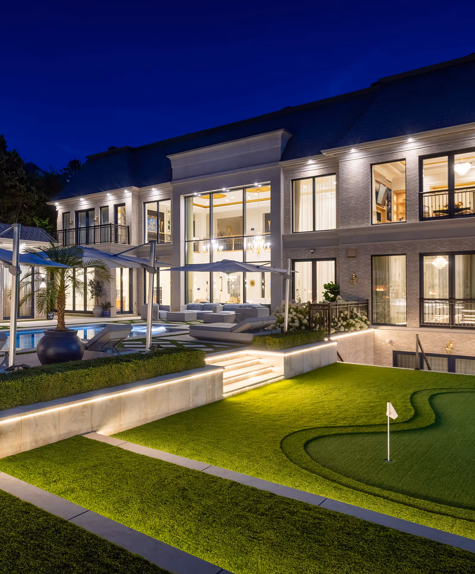 Night view of a luxury home with illuminated patio, pool, outdoor seating, and a putting green.