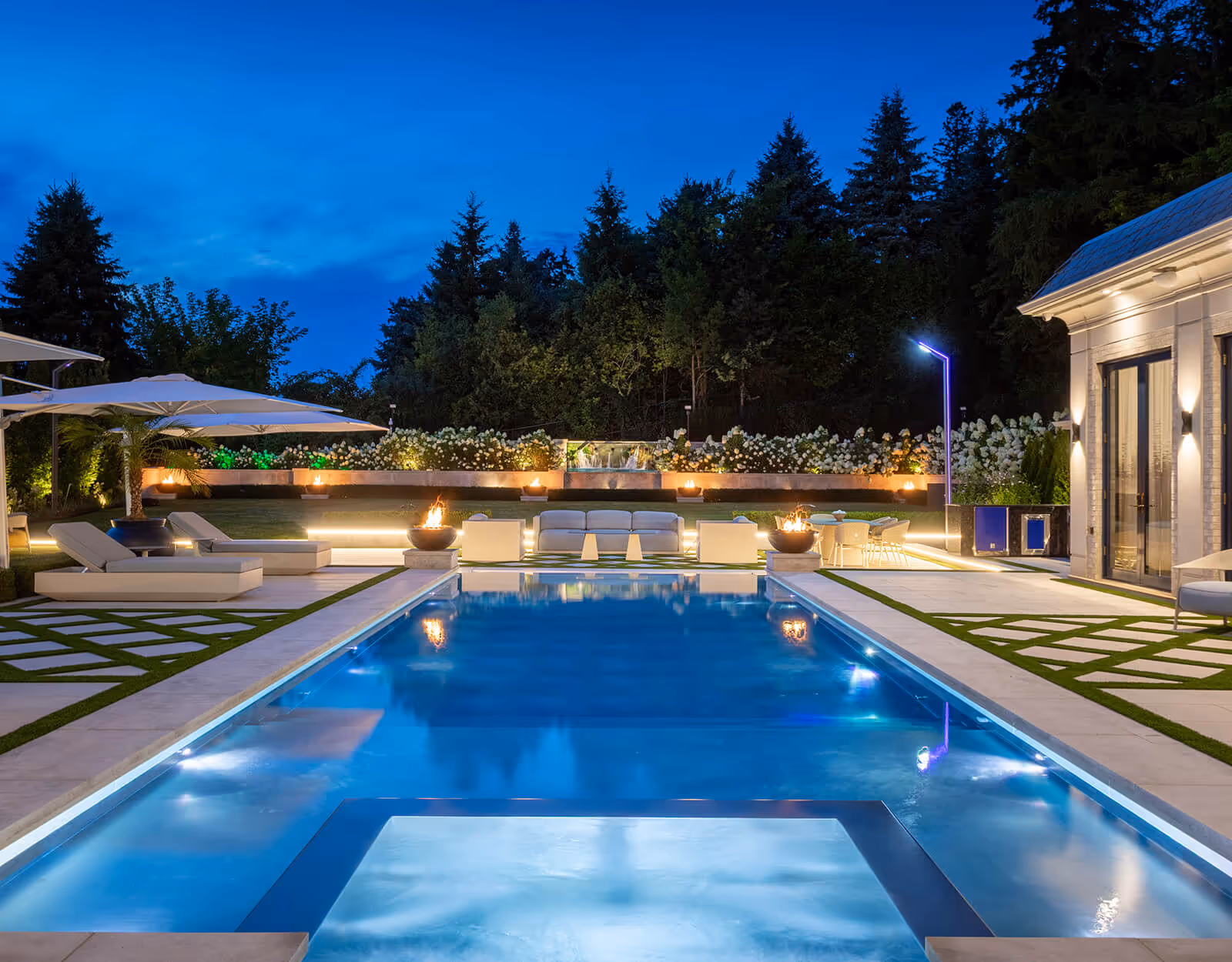 Luxurious backyard pool area at night with lounge chairs, fire pits, and illuminated seating.