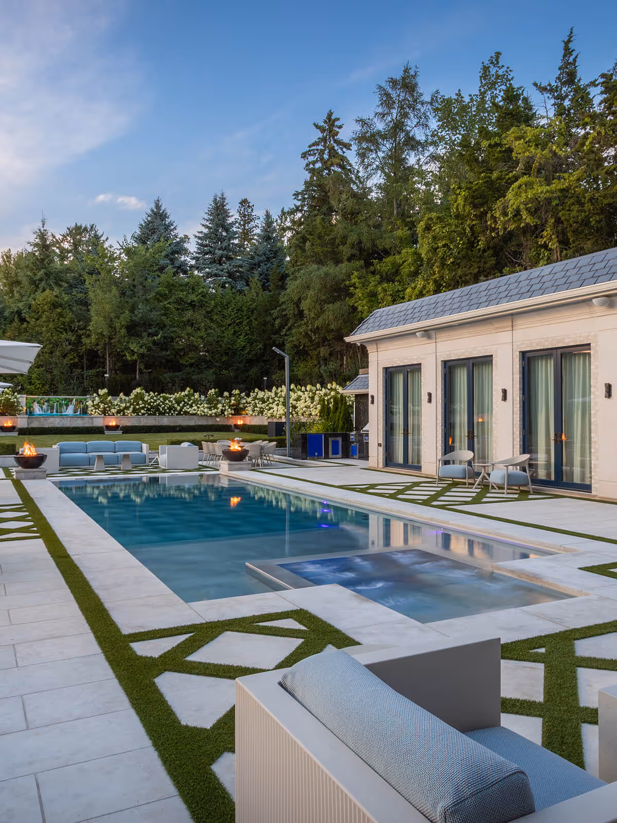 Modern backyard with a pool, spa, fire pits, outdoor seating, and surrounded by trees and flowers.