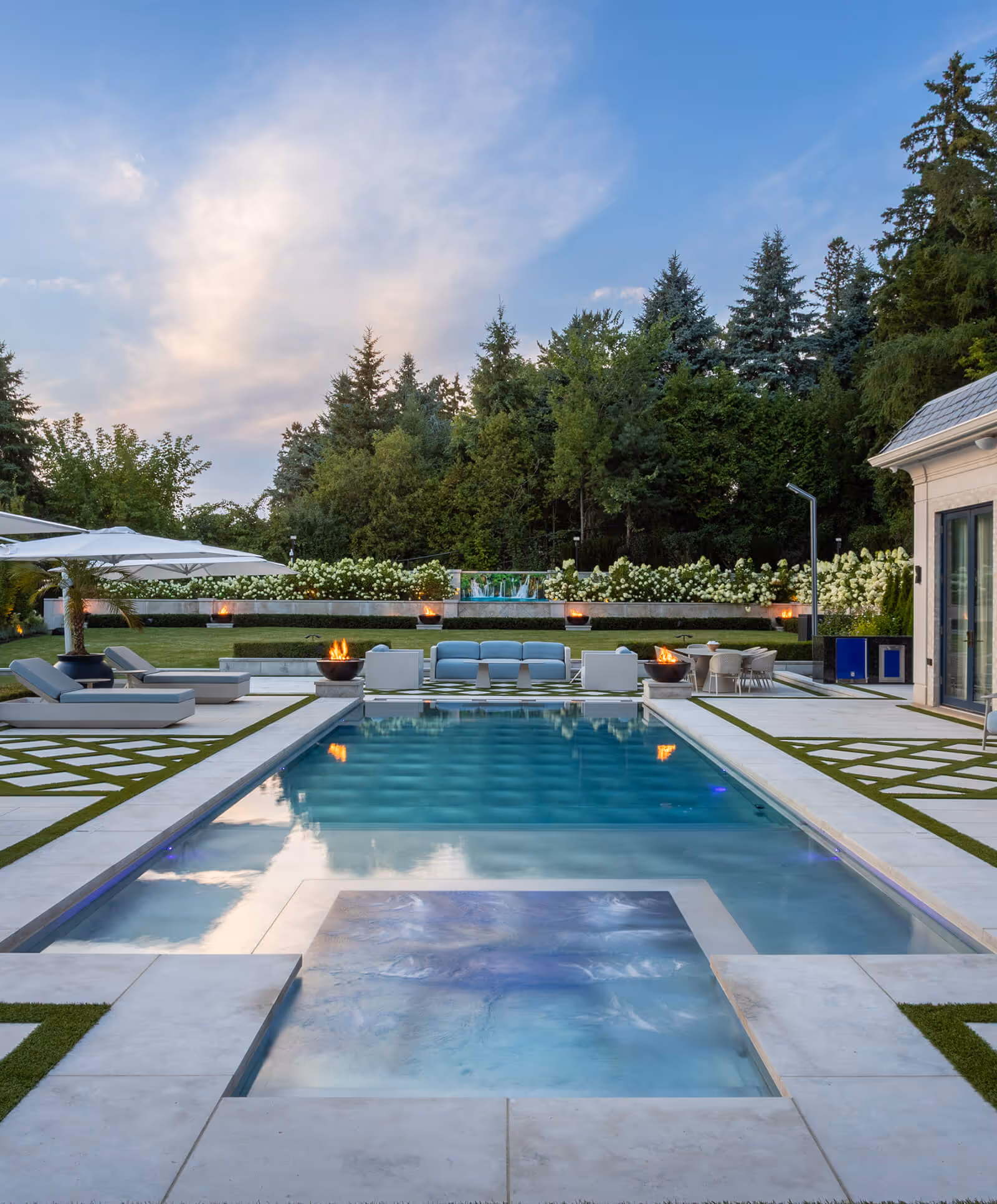 Modern outdoor swimming pool with hot tub, lounge chairs, fire pits, and seating area surrounded by greenery.