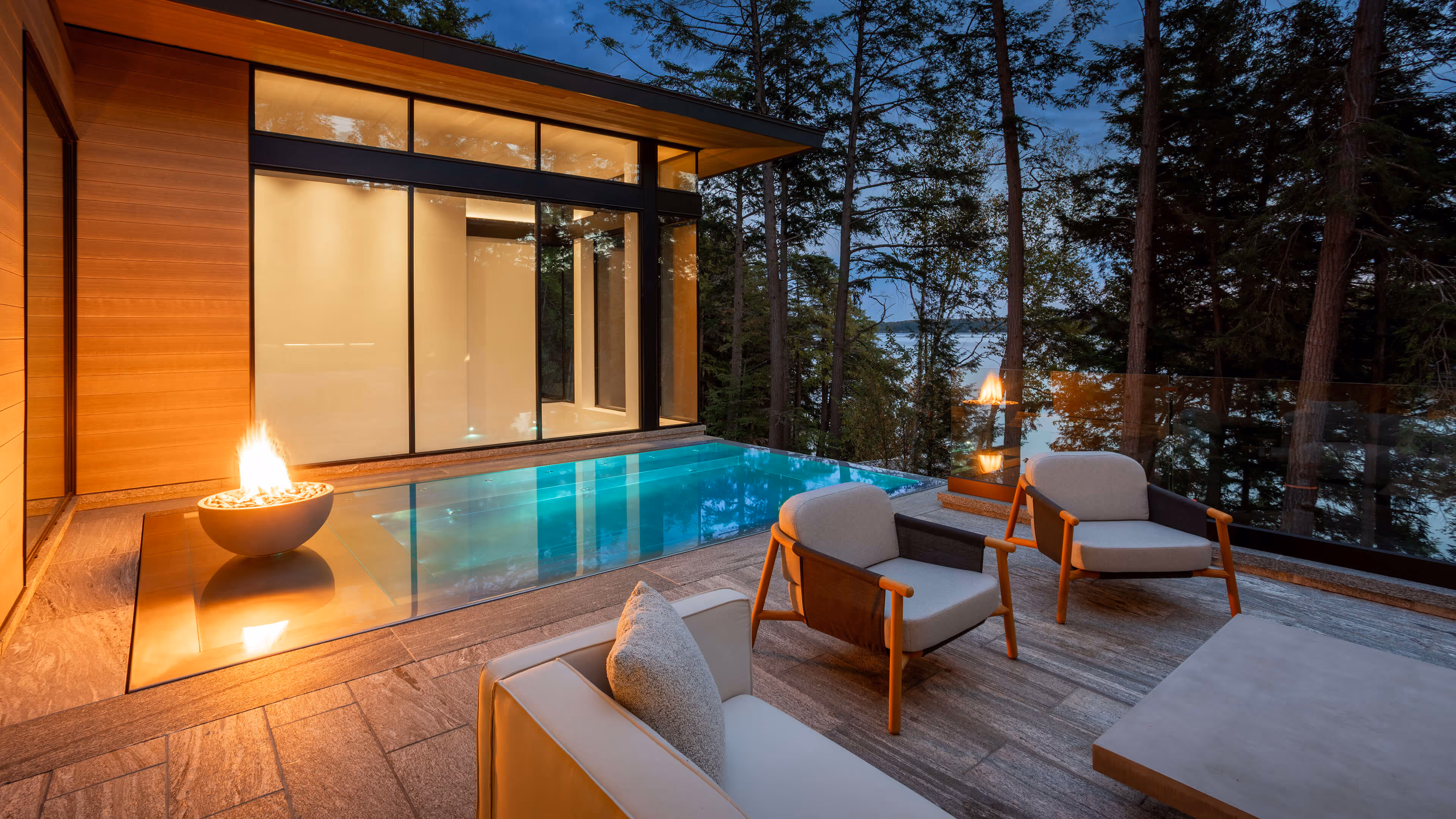 Modern patio with pool, fire pits, chairs, and trees overlooking a lake at dusk.