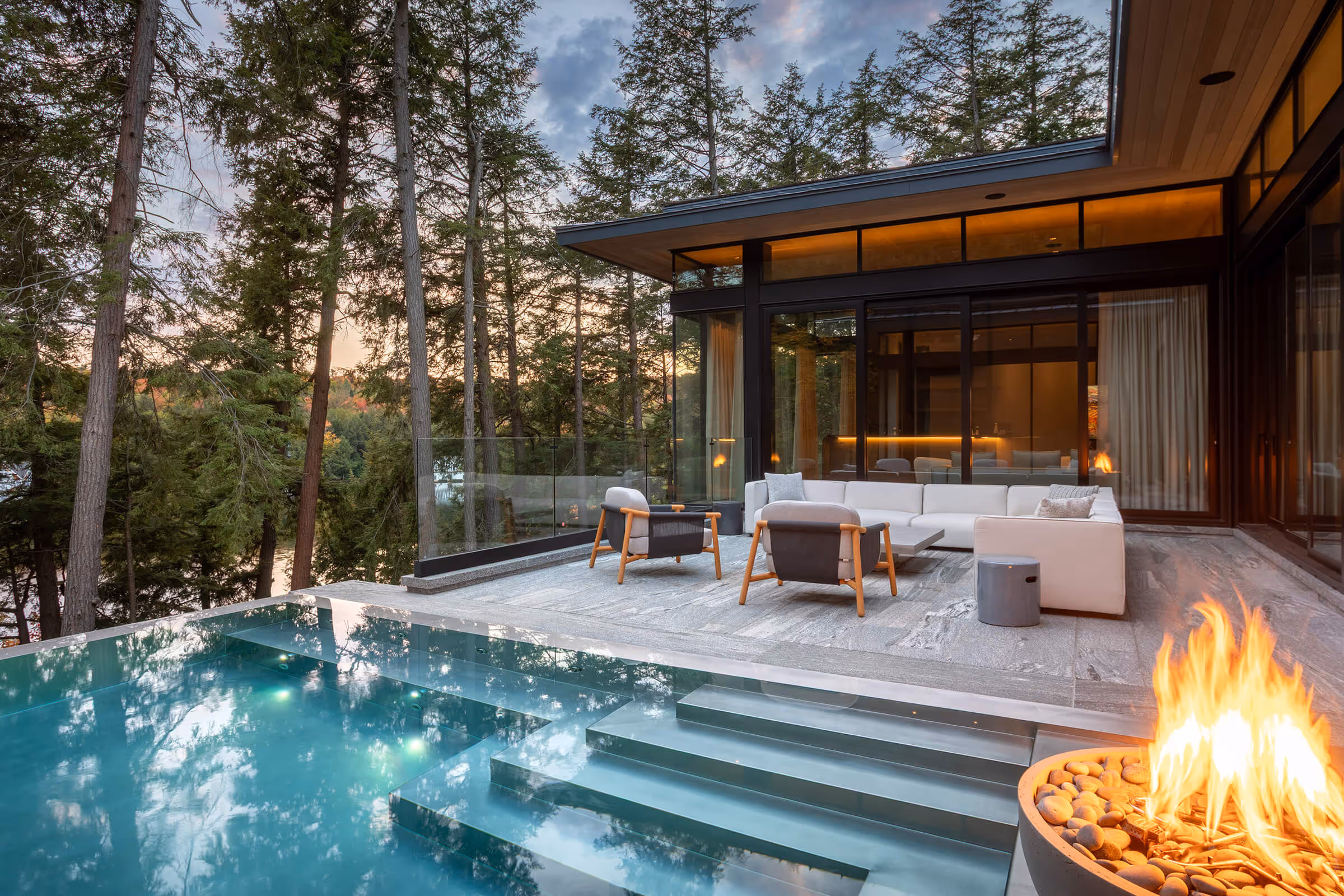 Modern patio with sofa, chairs, fire pit, and pool surrounded by tall trees at dusk.