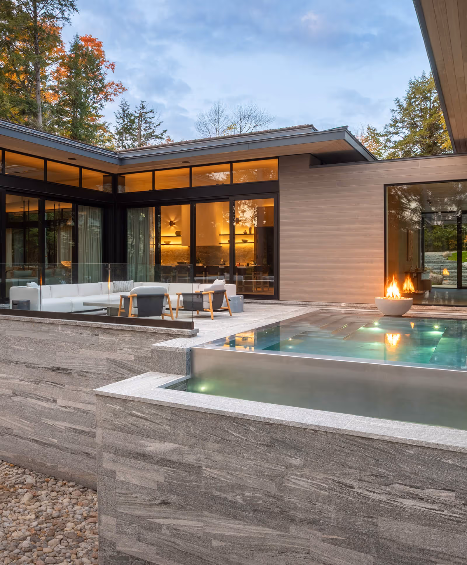 Modern outdoor patio with white sofa, chairs, glass fire pit, and an illuminated pool at dusk.
