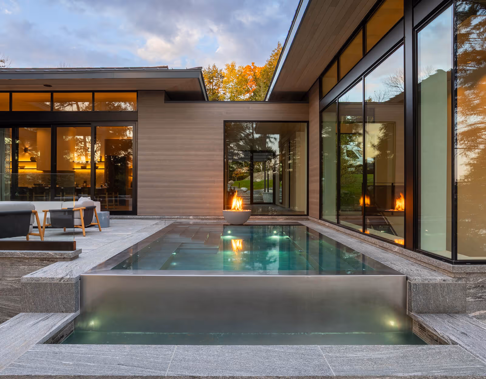 Modern backyard with an infinity-edge pool, a fire bowl, and seating near a house with large glass windows.