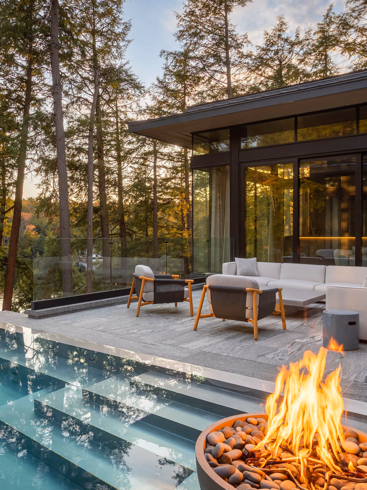 Modern patio with stone fire pit, glass railing, pool, lounge chairs, sofa, and forest view at sunset.