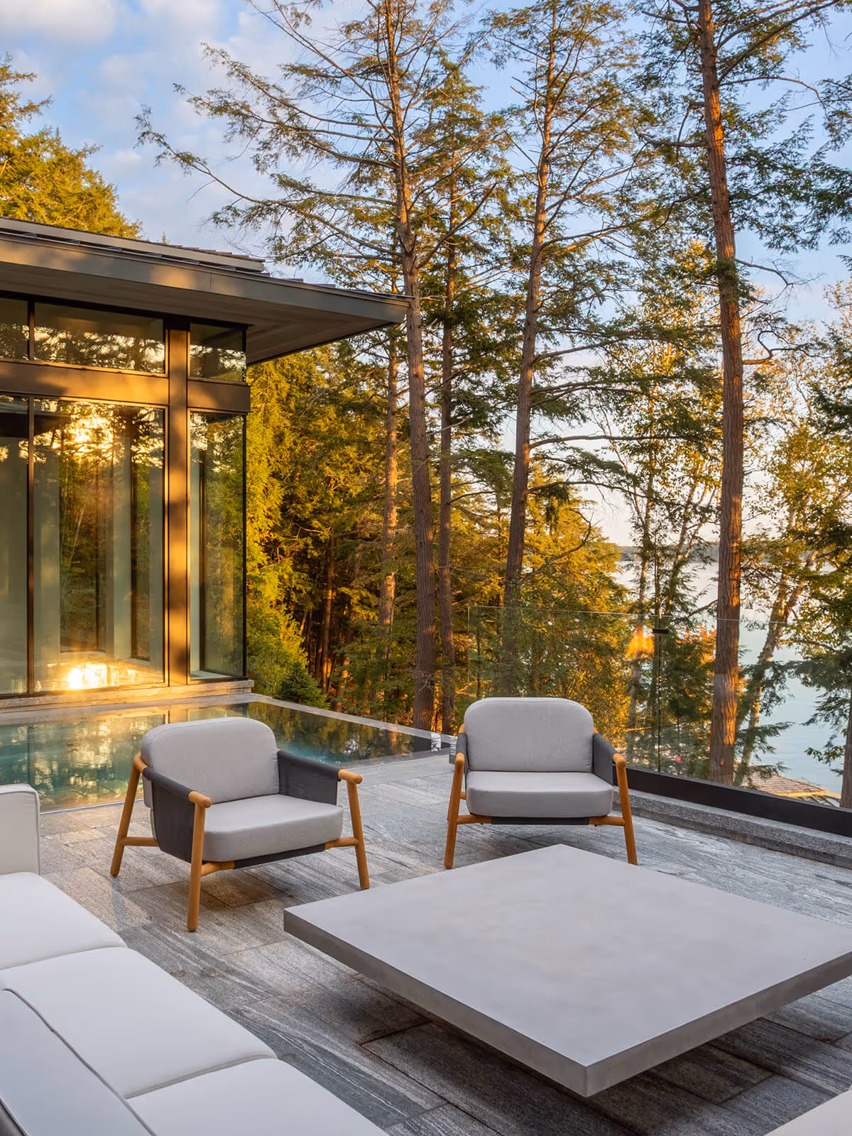 Modern patio seating with gray chairs and sofa near a glass-walled house overlooking trees and water.