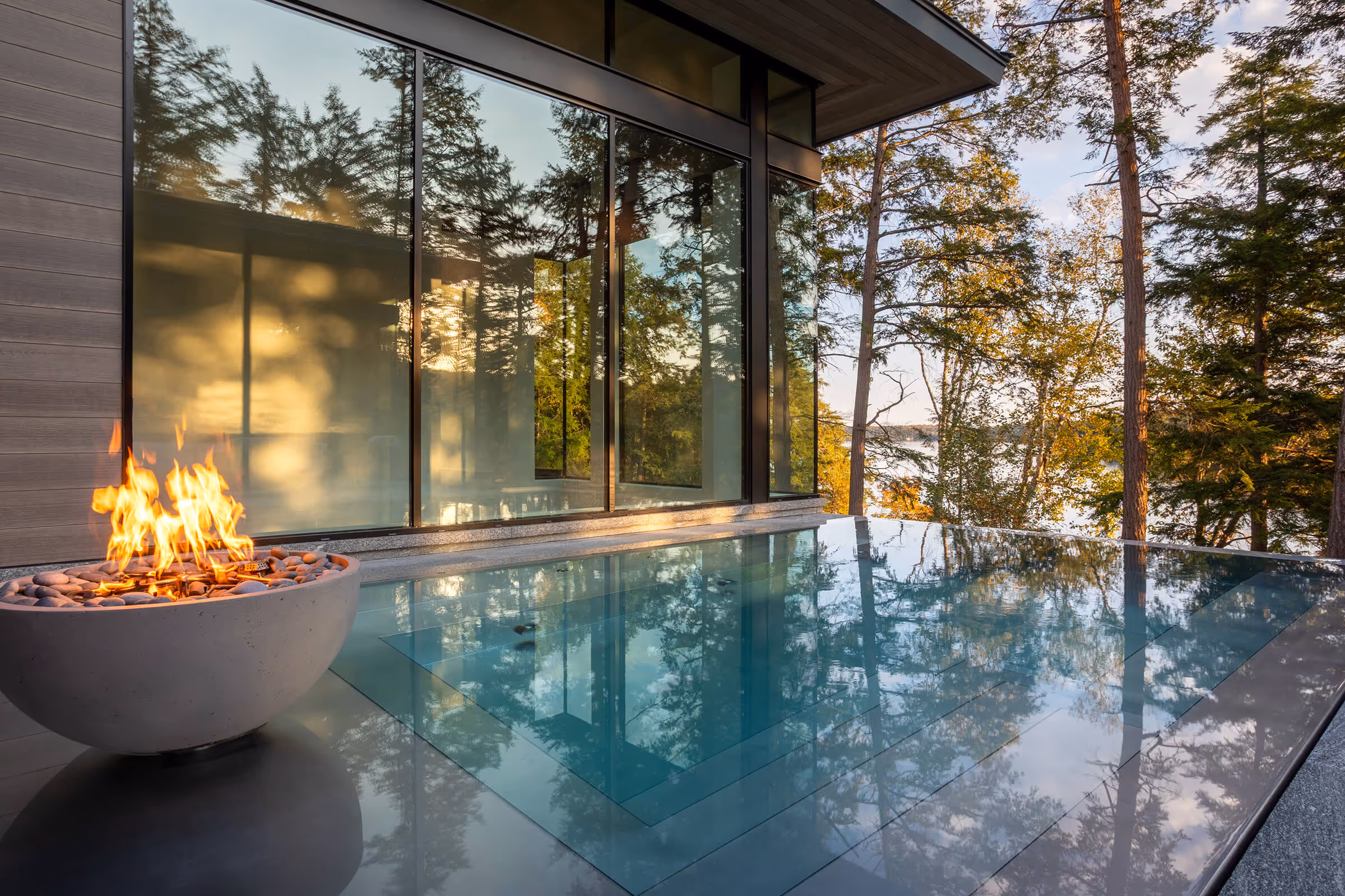 Modern house patio with glass pool reflecting trees and a fire bowl with flames at sunset.