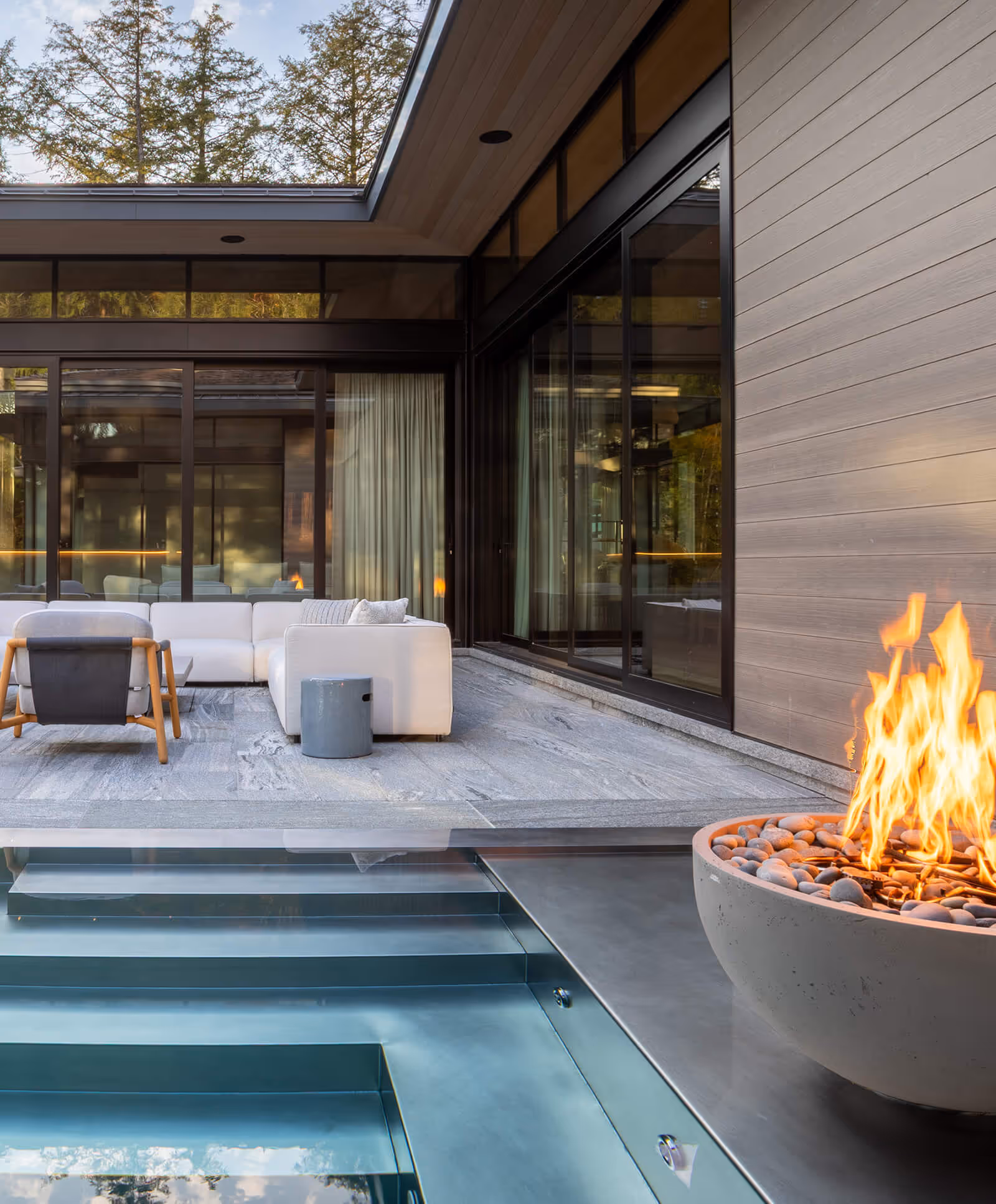Modern outdoor patio with white couches, glass walls, hot fire bowl, and pool steps.
