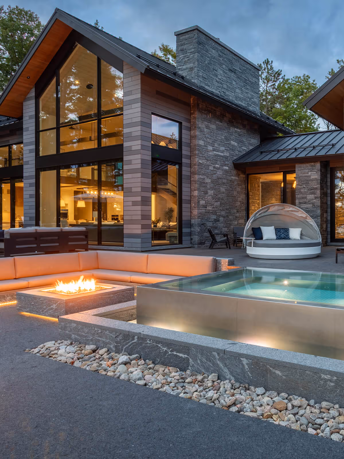 Modern house patio at dusk with fire pit, sunken seating, hot tub, and round outdoor daybed.