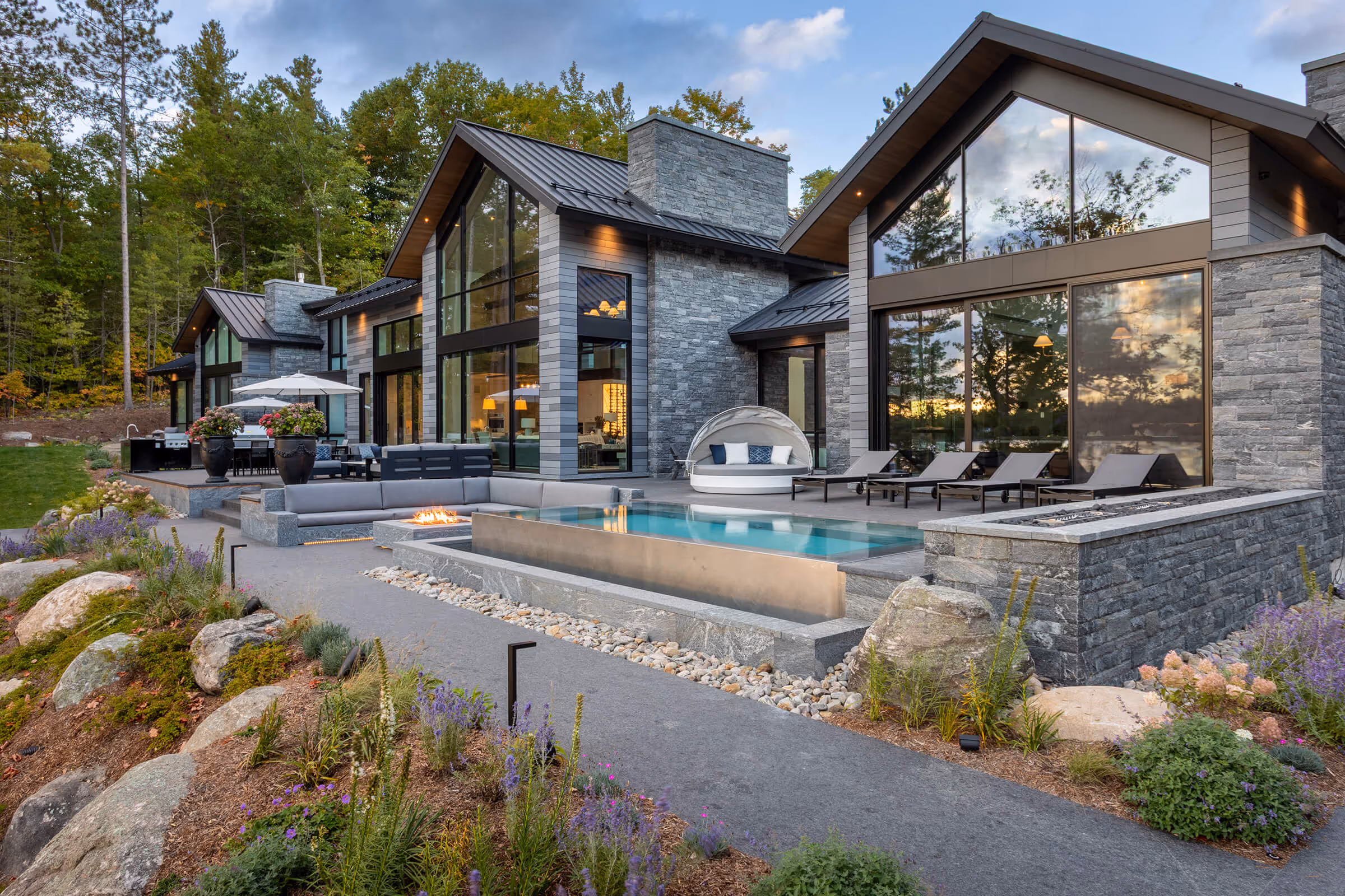 Modern stone house with large windows, patio pool, lounge chairs, outdoor fire pit, and garden pathway.