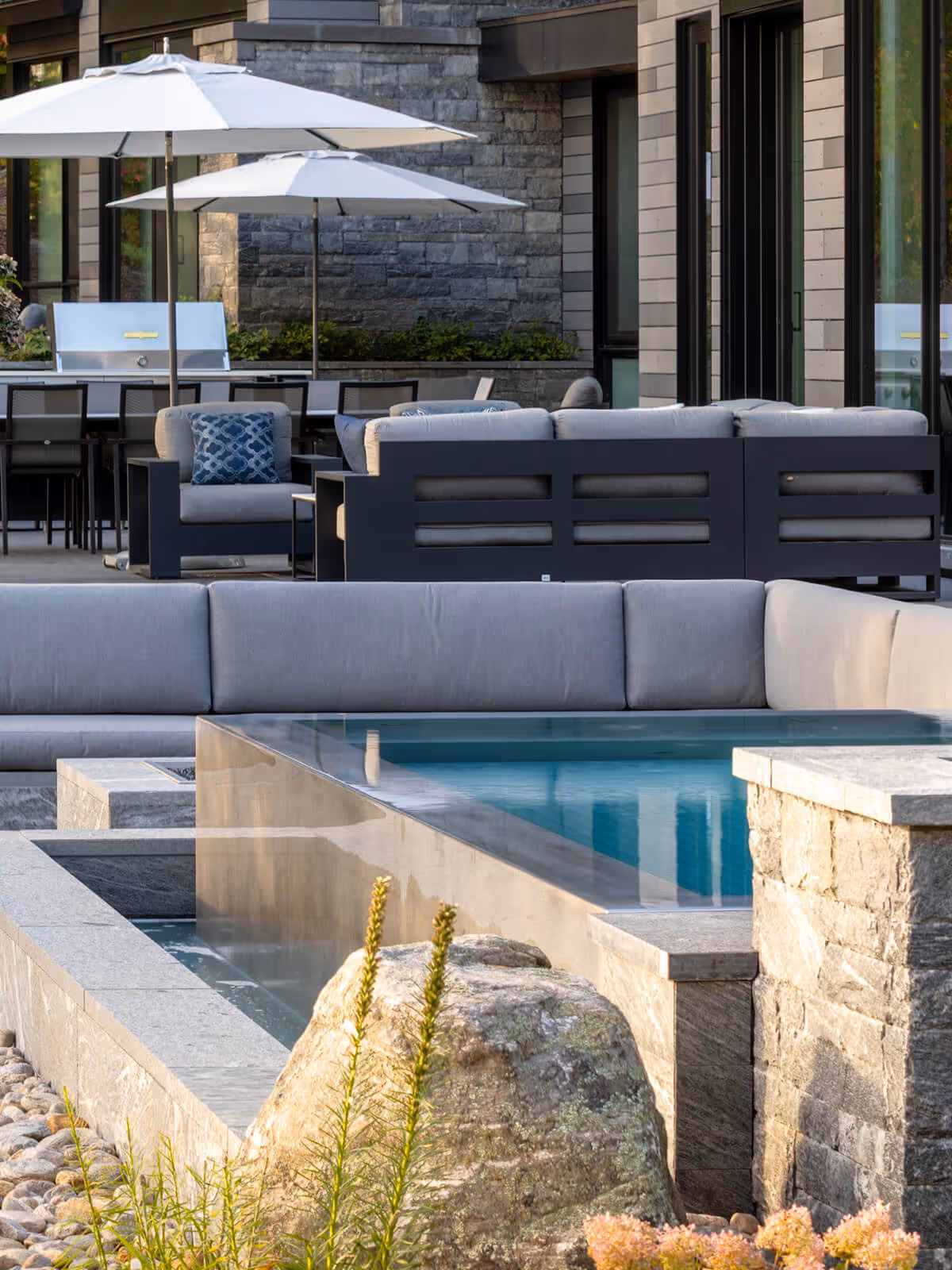 Modern outdoor seating area with gray sofas, umbrellas, stone firepit, and water features.