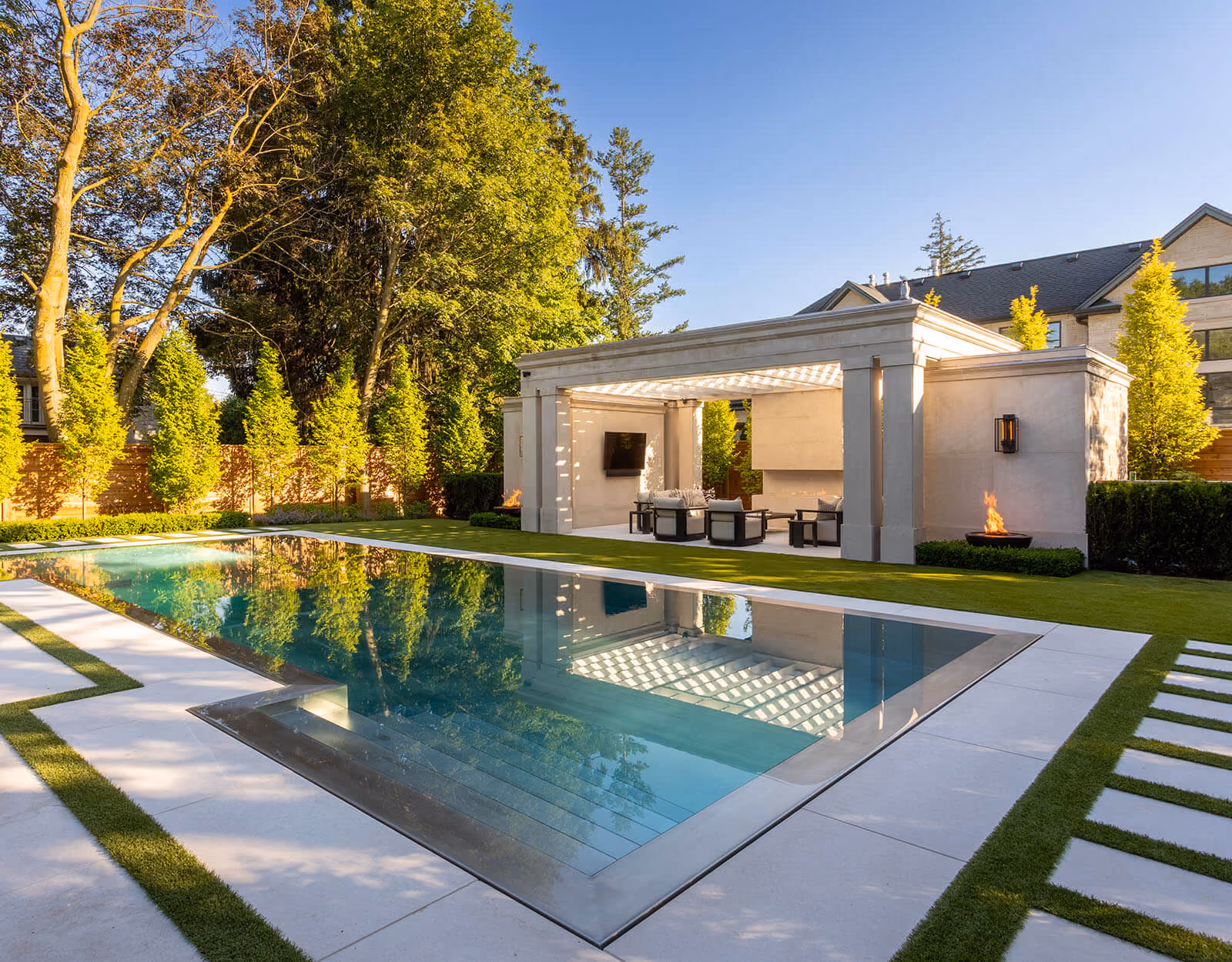 Modern outdoor pool beside a covered patio with seating, fire features, and a TV in a sunny backyard.