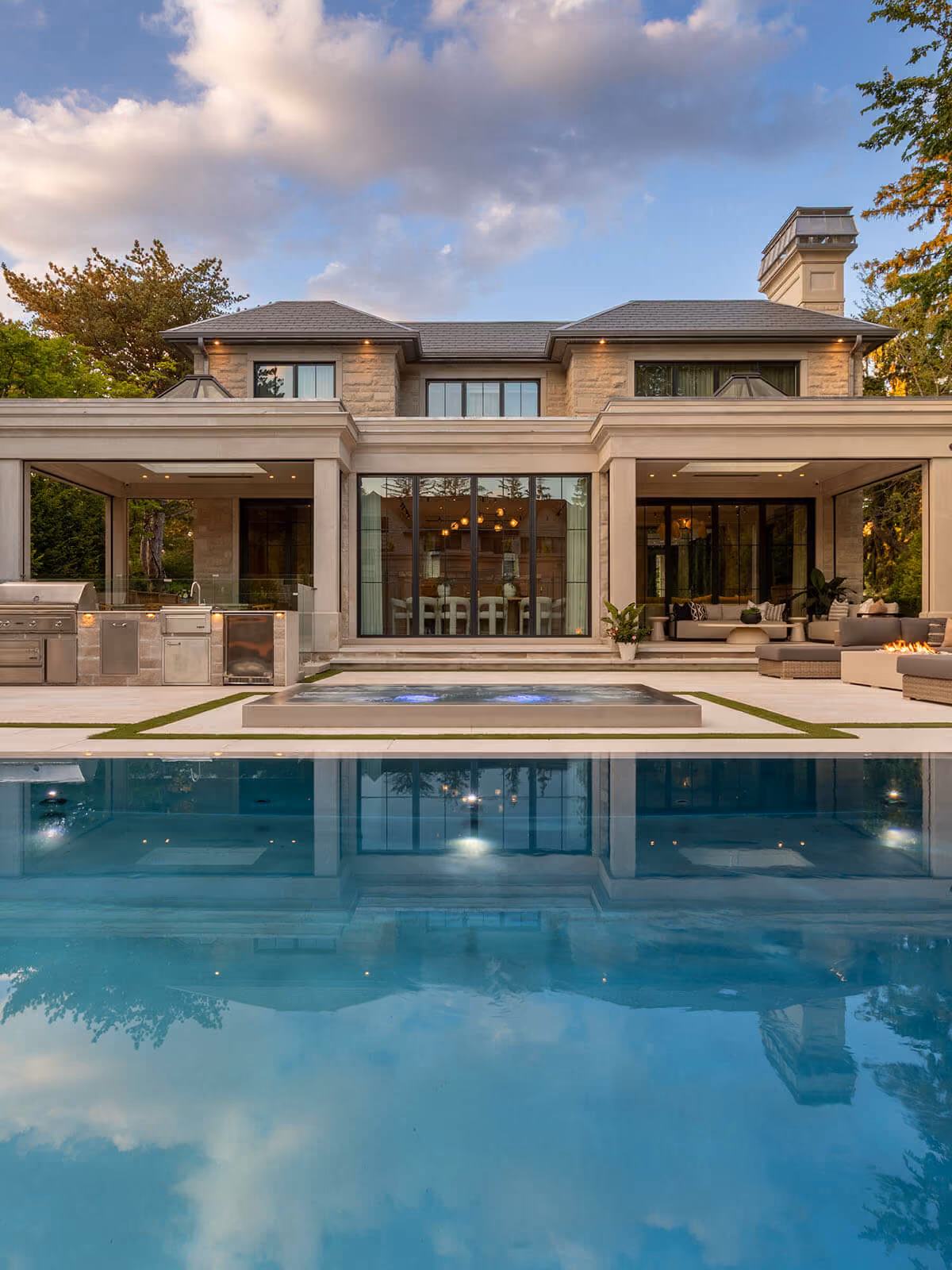 Modern two-story house with large windows, outdoor kitchen, seating area, hot tub, and pool.