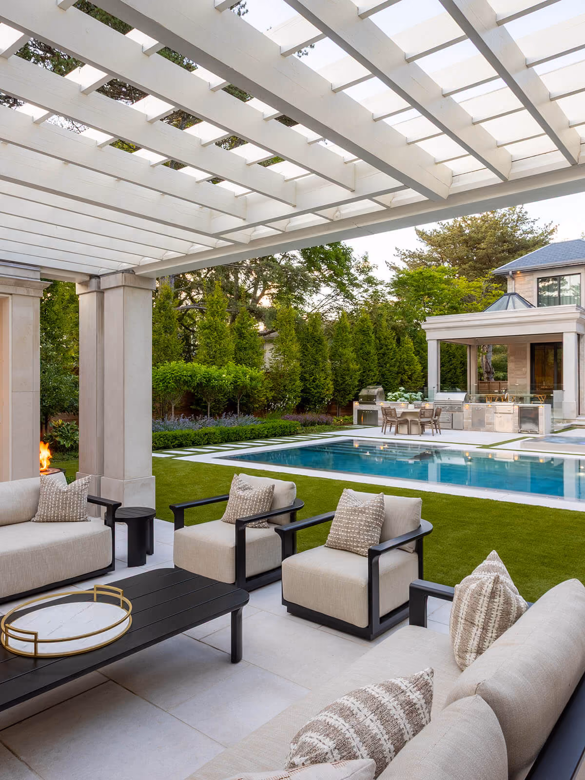Modern outdoor patio with beige cushioned sofas, black coffee table, lawn, pool, and outdoor kitchen area.