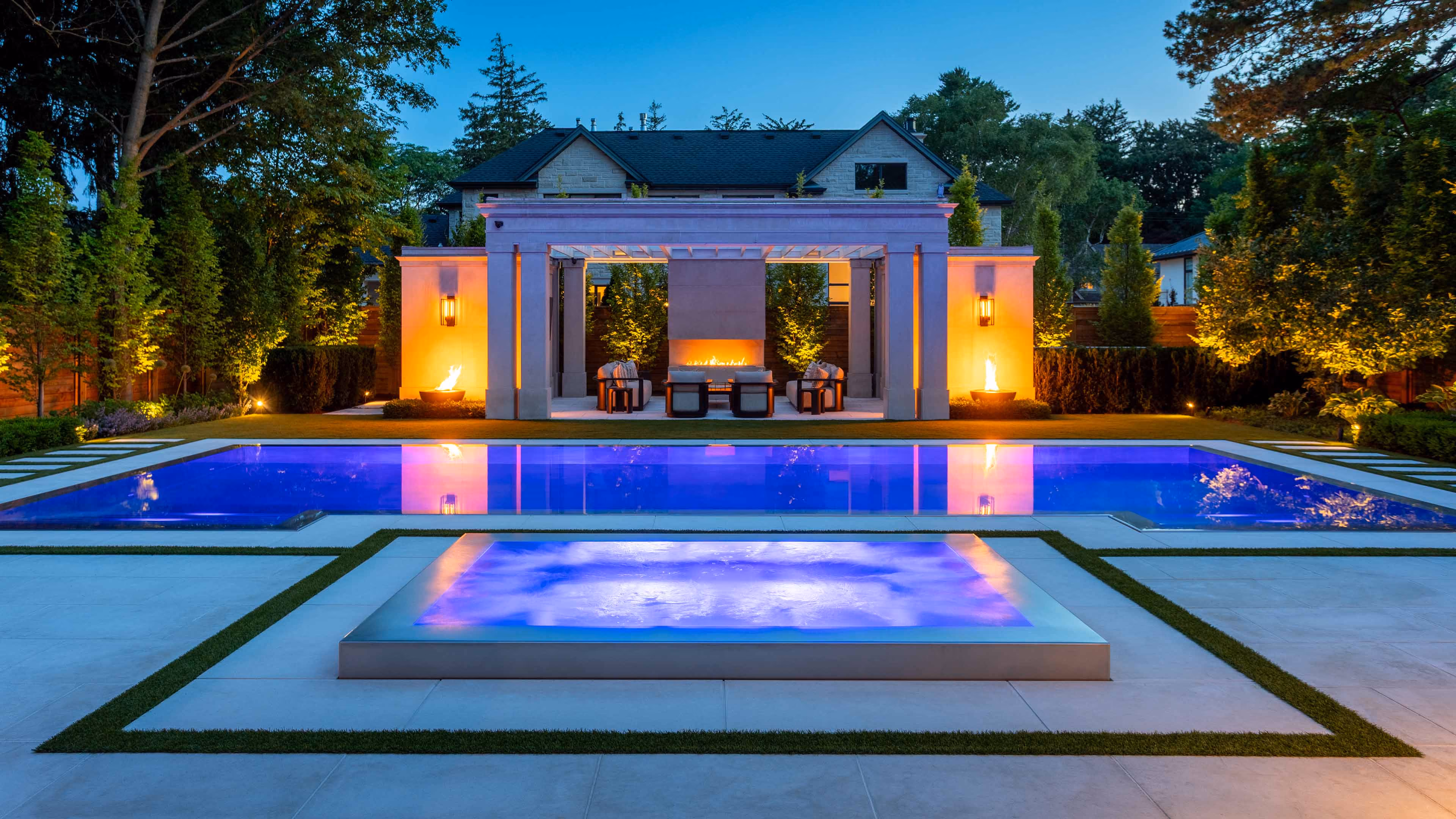 Modern luxury home with lit outdoor pool, hot tub, lounge seating, and outdoor kitchen at dusk.
